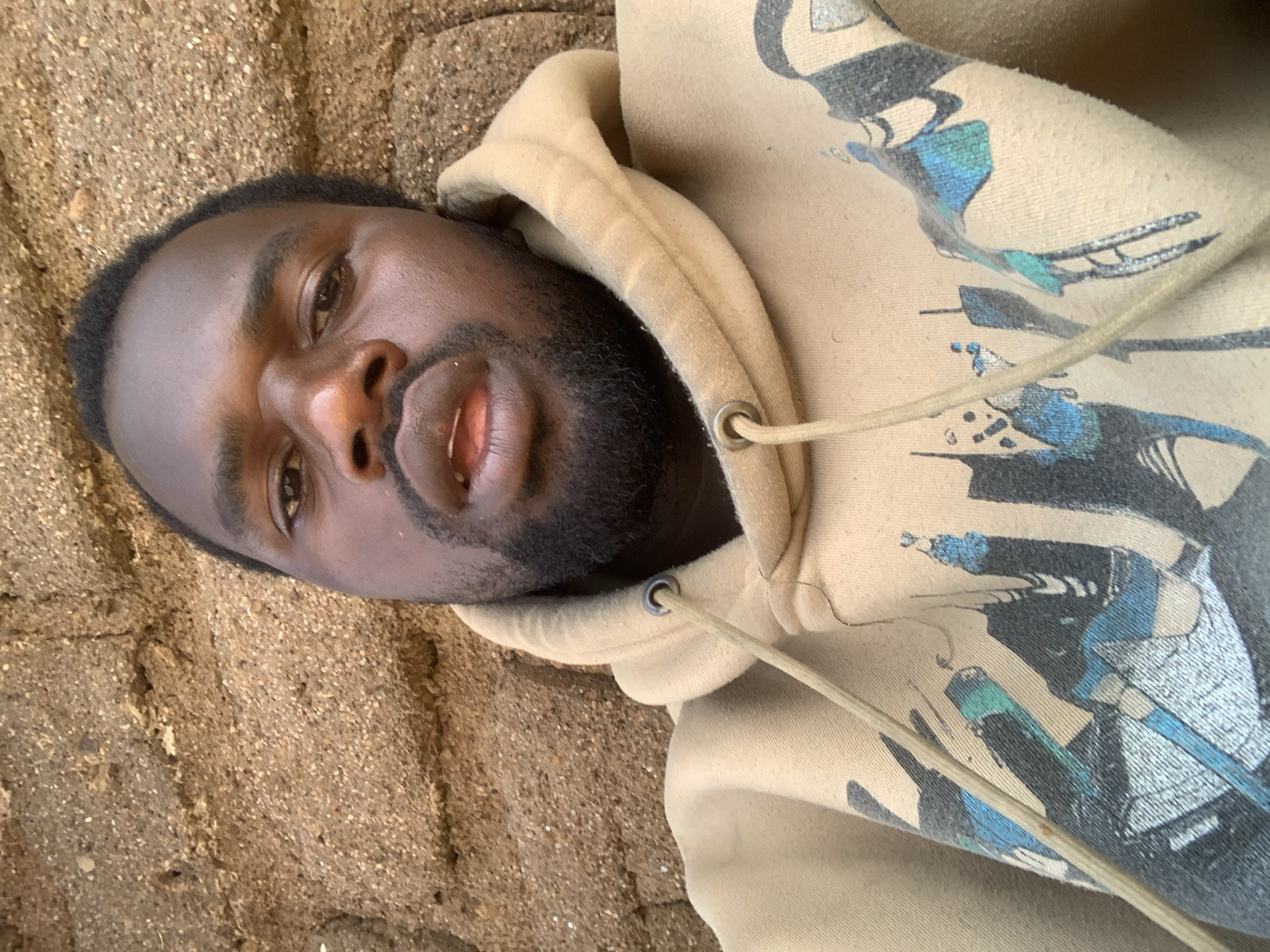 A medium shot photograph shows a Black man in a tan hoodie against a textured, light brown brick wall. The man is positioned centrally in the frame, facing the camera directly with a neutral expression. His head is slightly tilted, and his eyes are open, looking forward. He has short, dark hair and a short beard and mustache. The hoodie is a light tan color with a graphic print on the front featuring stylized black and blue imagery. A light tan drawstring with a metal eyelet is visible on the left side of the hoodie. The brick wall behind him is rough and uneven, with visible individual stones and mortar. The lighting suggests an outdoor environment. The specific location of Buga da Gindi, Nigeria is not ascertainable from the image itself.