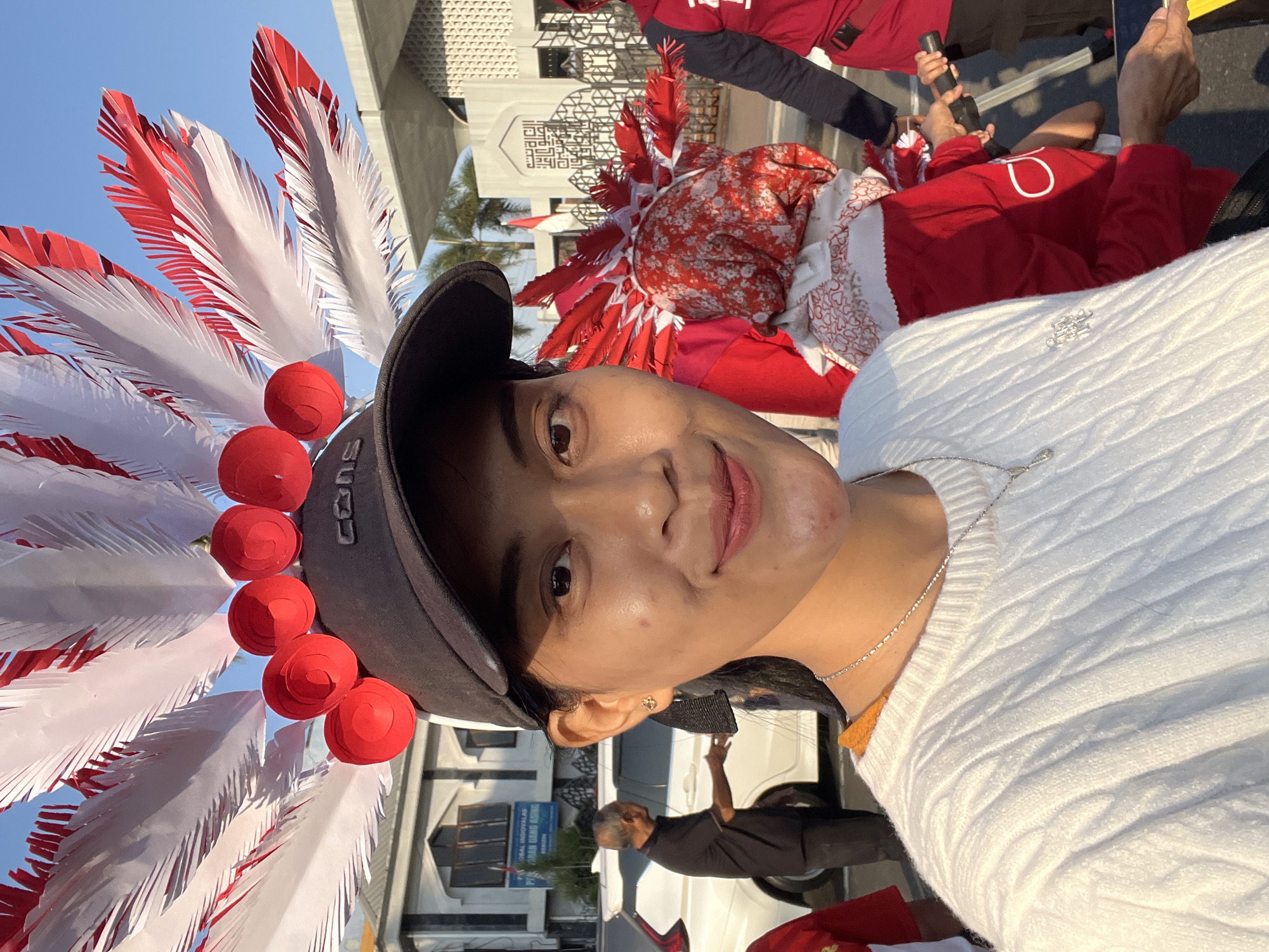 In Kecamatan Pati, Indonesia, a young woman wearing a black baseball cap adorned with red and white feathered headwear takes a selfie. She is dressed in a white cable-knit sweater and a silver necklace. Behind her, a man in a black shirt gestures towards a white car. Further in the background, another person wears a red and white patterned headscarf and a red garment, participating in a festive or celebratory event, possibly a parade, indicated by the numerous red and white decorations resembling feathers and circular ornaments. A building with Islamic architectural elements is visible behind the participants. The scene takes place outdoors during daylight, under a clear blue sky.