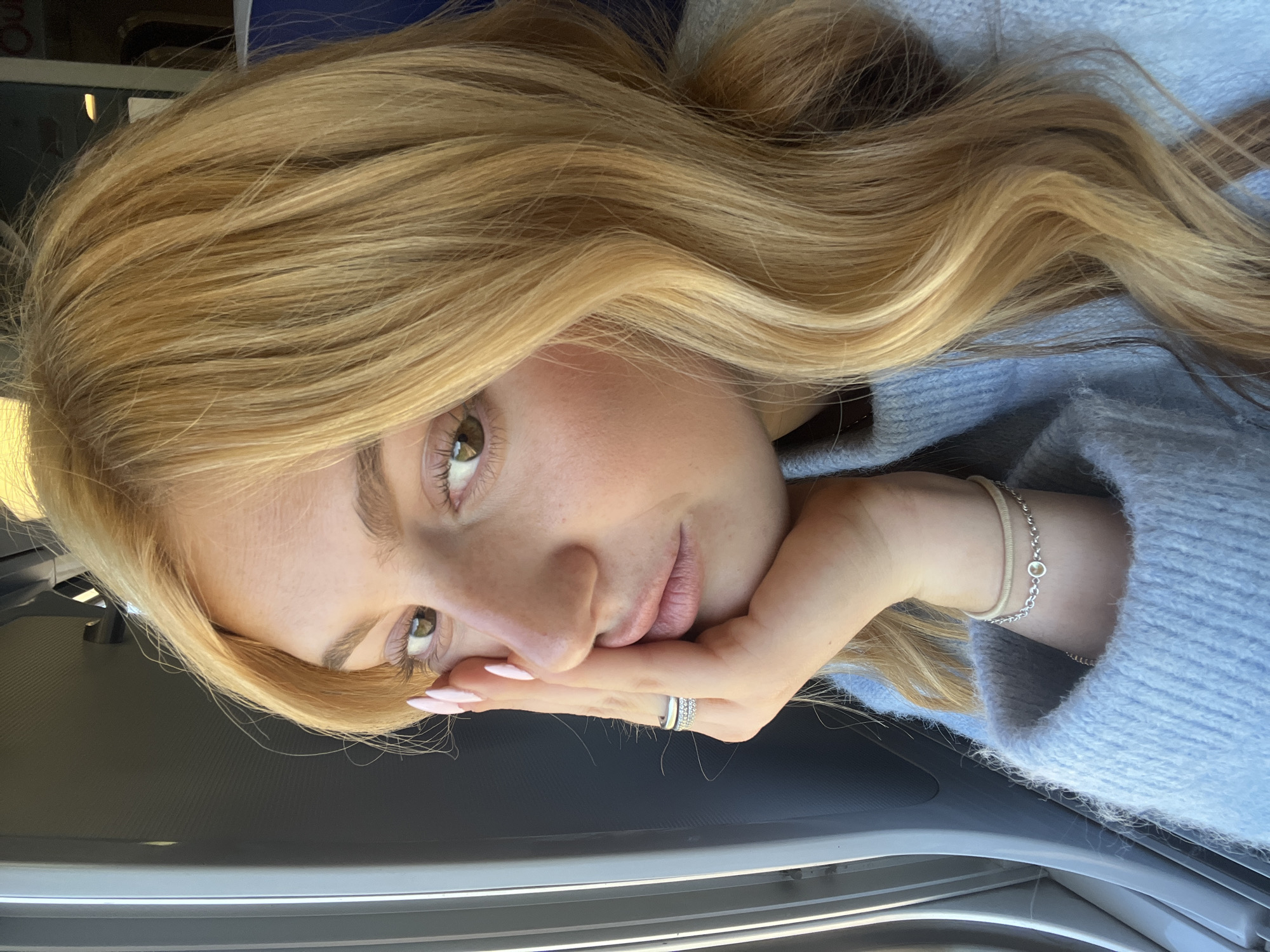 A close-up selfie shows a young woman with long, wavy blonde hair with lighter highlights, resting her chin on her right hand. Her head is tilted, giving the impression of lying down or leaning significantly. She has green or hazel eyes with visible eyelashes and a light complexion with subtle freckles. Her expression is calm and direct. She is wearing a light blue knitted sweater. On her right hand, she has light pink manicured nails and two rings on her ring finger – one stacked with small stones and another plain band. A silver chain bracelet with a small, round yellow charm and a beige hair tie are visible on her right wrist. The background is dark and indistinct, featuring grey and metallic linear elements that suggest the interior of a vehicle, possibly a car door or dashboard. Bright natural light illuminates her face from the left side, indicating it is daytime. No text is visible. The scene is set in Sarry, France.