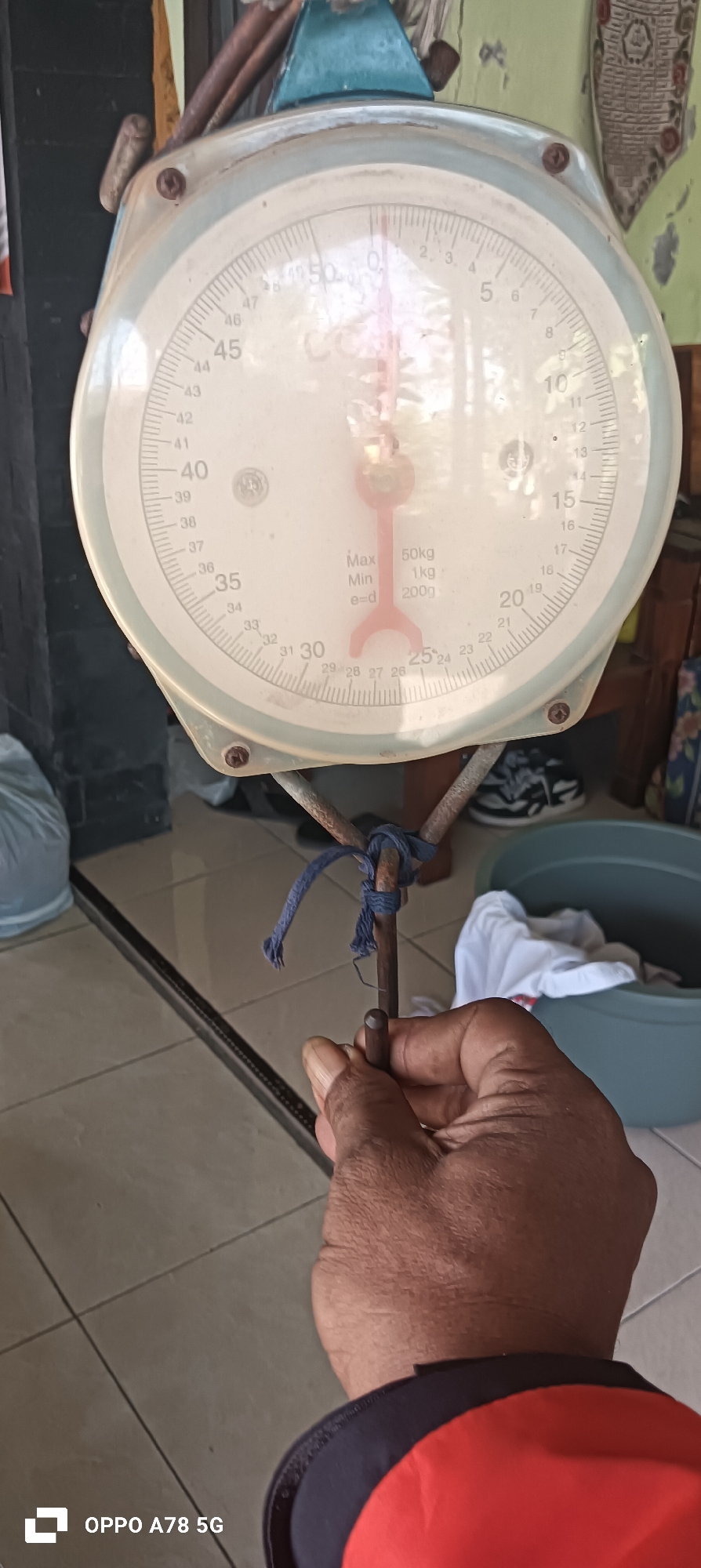 A close-up shot focuses on a hand holding the hook of a vintage mechanical scale, with the scale's dial and face prominently displayed. The scale, likely located in Buncitan, Indonesia, shows markings up to 50 kg, with a needle pointing slightly above the 25 kg mark. The dial has various measurements and indicators, including "Max 50kg", "Min 1kg", and "e=d 200g". The scale is suspended by rusty metal rods tied with a blue string. The background reveals a tiled floor and partially visible objects like a blue tub, white fabric, and a dark wall. The person's hand, with visible skin texture and short fingernails, is holding the hook. The overall scene suggests an everyday setting, possibly a shop or a household, where the scale is in use.