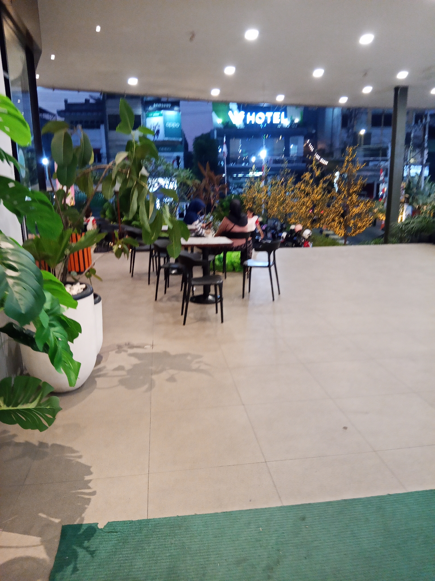 The image depicts an outdoor seating area at dusk, possibly a cafe or restaurant. In the foreground, a large white planter holds a vibrant green monstera plant, casting a shadow on the tiled floor. A dark green mat runs along the bottom edge of the frame. In the mid-ground, several black tables and chairs are arranged, with a few patrons seated at one of the tables, engaged in conversation or eating.
In the background, a brightly lit sign reads "W HOTEL," indicating a hotel establishment. Further in the distance, a large, decorated tree, adorned with numerous small yellow lights, suggests a festive atmosphere, possibly related to a holiday season. The environment appears to be urban, with buildings and streetlights visible beyond the decorated tree. The lighting is dim, consistent with evening hours, and the overall impression is one of a public space with people enjoying a meal or drinks in a relaxed setting.
While the image does not explicitly state the location, "W HOTEL" is a global hotel brand with locations in many cities. To pinpoint the specific location in Lubuklinggau, Indonesia, further information would be required.