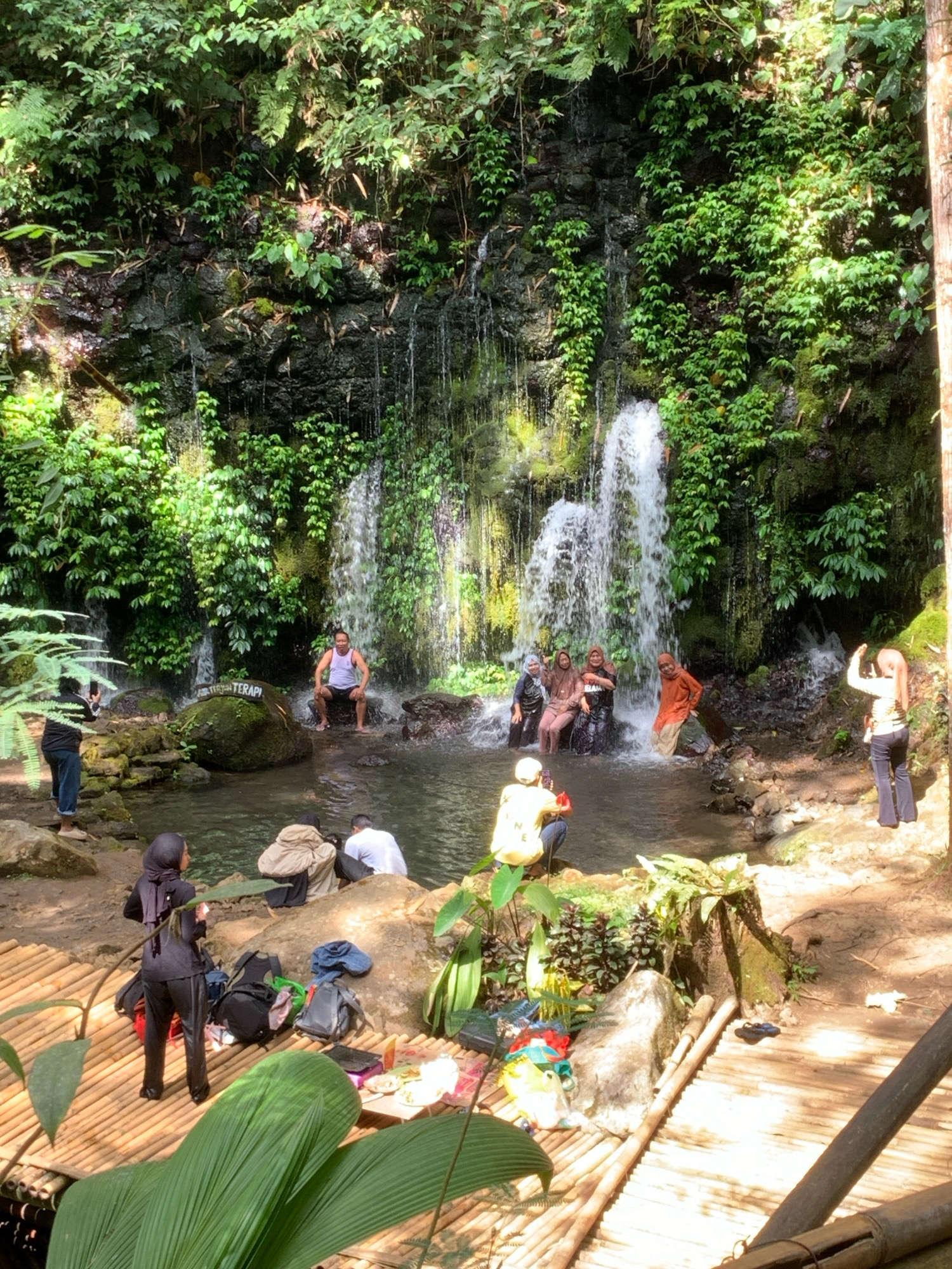 This is a vertical, eye-level, medium-wide shot of a waterfall cascading down a mossy, rocky cliff face covered in lush green foliage. A group of people are interacting with the waterfall and surrounding area. In the pool at the base of the waterfall, a man is sitting on a rock, with several women in headscarves wading or sitting in the water. To the left of the pool, a person in a black jacket and jeans is taking a photo of the waterfall with their phone. Further back, near the edge of the pool, are more people, some sitting and some standing. In the foreground, a person in a black hijab and pants is standing on a bamboo platform, with several backpacks and bags nearby. Another person in a yellow shirt is kneeling on the bamboo platform, holding a camera. The ground is a mix of dirt and rocks, with more bamboo structures. The overall scene depicts a natural tourist attraction, likely a popular spot for recreation and photography. This scene is located in Pronojiwo, Indonesia.