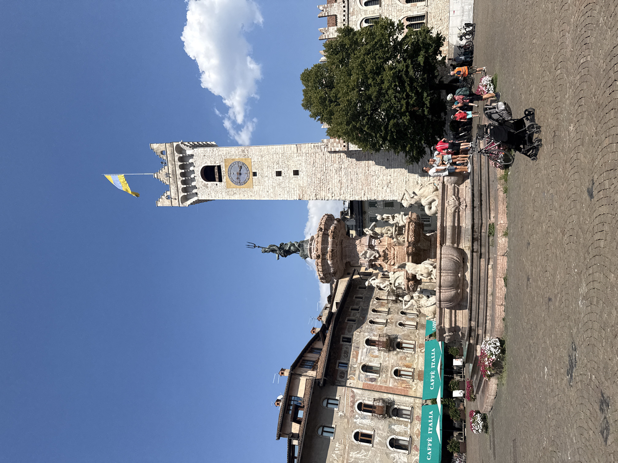 The image shows a wide-angle view of Piazza Duomo in Trento, Italy, on a sunny day. In the center of the piazza is the Fountain of Neptune, a Baroque masterpiece featuring Neptune with his trident at the top, surrounded by mythological figures and sea creatures. To the right of the fountain stands the Torre Civica, a tall, pale stone clock tower with a yellow and blue flag flying from its top. The clock face shows the time as approximately 11:10. The buildings surrounding the piazza have historical architecture with painted facades. Green awnings with the text "CAFFÈ ITALIA" are visible on the ground floor of one building. In the foreground, a cobblestone pavement is visible. Several people are present in the piazza, including a group standing near the fountain, individuals with bicycles, and a person pushing a stroller. The sky is a clear blue with a few white clouds. The overall scene depicts a lively public square in an Italian city, likely during daytime leisure activities.