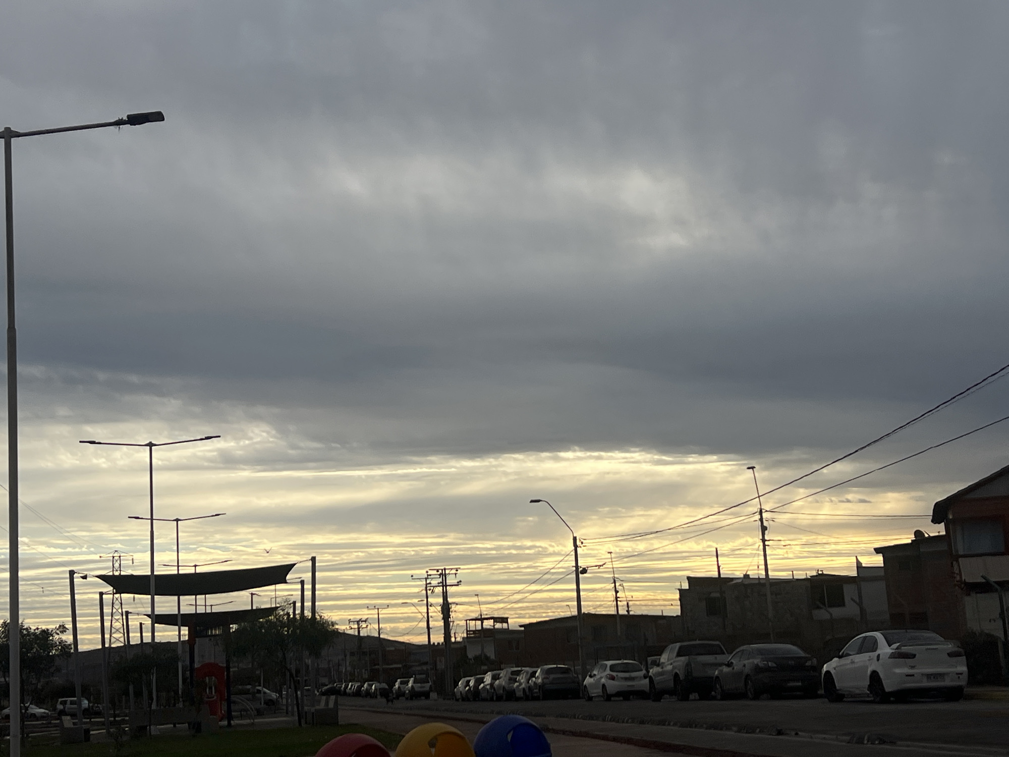 The image depicts a street scene in Arica, Chile, under an overcast sky with streaks of yellow and orange light from a sunset or sunrise. A prominent street lamp stands on the left side of the frame, with a dark, rectangular lamp head. In the middle ground, a park-like area features a large, abstract sculpture resembling a ship or sails, with multiple horizontal poles. Behind the sculpture, more street lamps and power lines are visible, along with trees. The foreground shows the edge of a street with several parked cars, including a white sedan, a dark sedan, and a truck. Further down the street, more cars are parked and some buildings are visible. The overall atmosphere is quiet, with no people clearly visible. The composition suggests an urban environment on the outskirts of the city, with a focus on the sky and street infrastructure.
