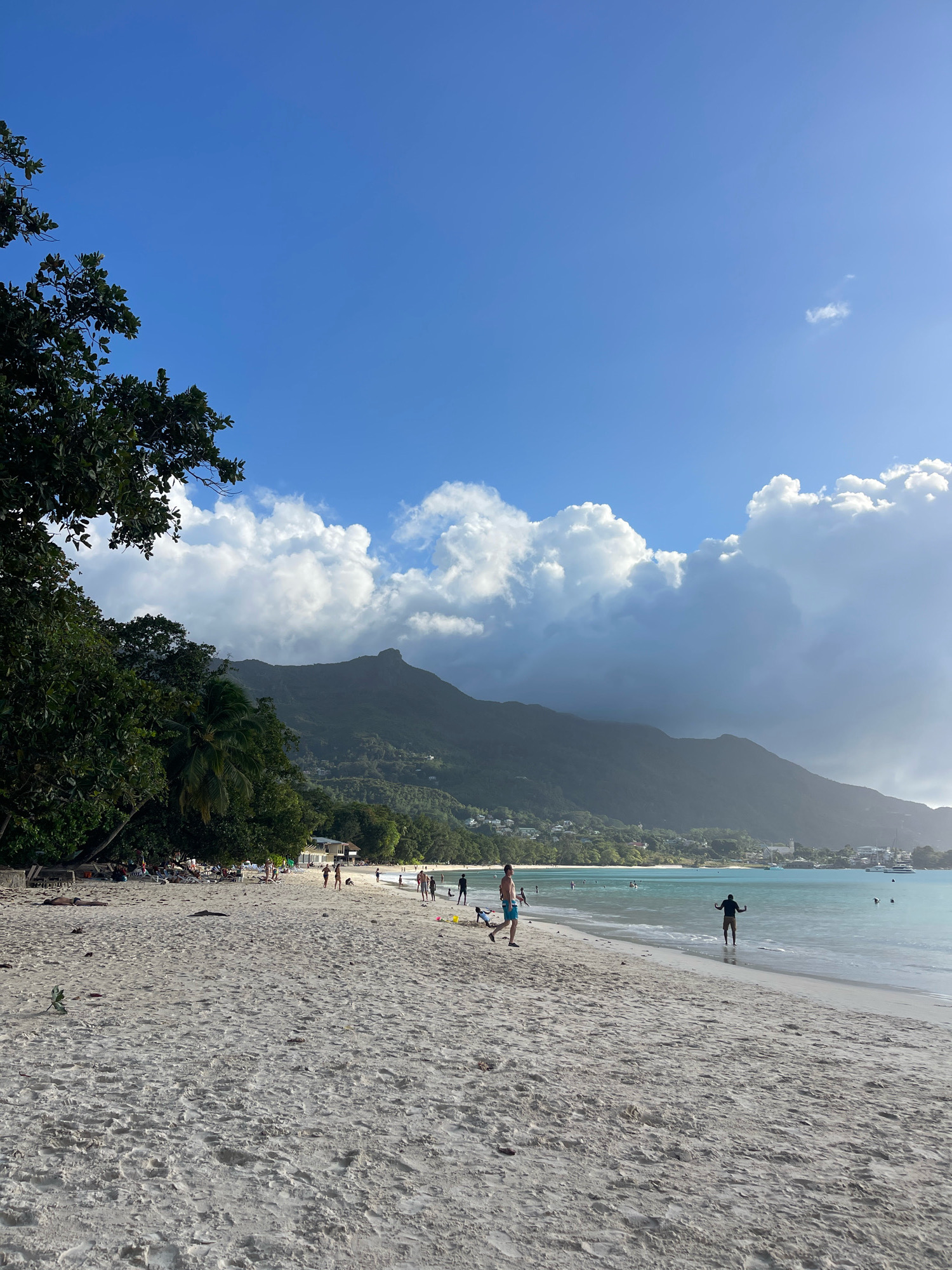 The image depicts a wide, sandy beach with turquoise water under a bright blue sky with scattered white clouds. Lush green trees line the left side of the beach, with some vegetation and buildings visible further down the coastline. In the background, a large, verdant mountain range dominates the horizon. Several people are scattered across the beach and in the shallow water, some walking, others wading, and a few appear to be swimming. A solitary figure stands in the water with arms outstretched, facing the camera. The sand is textured with footprints and small debris. The overall scene suggests a tropical, leisurely day at the beach.