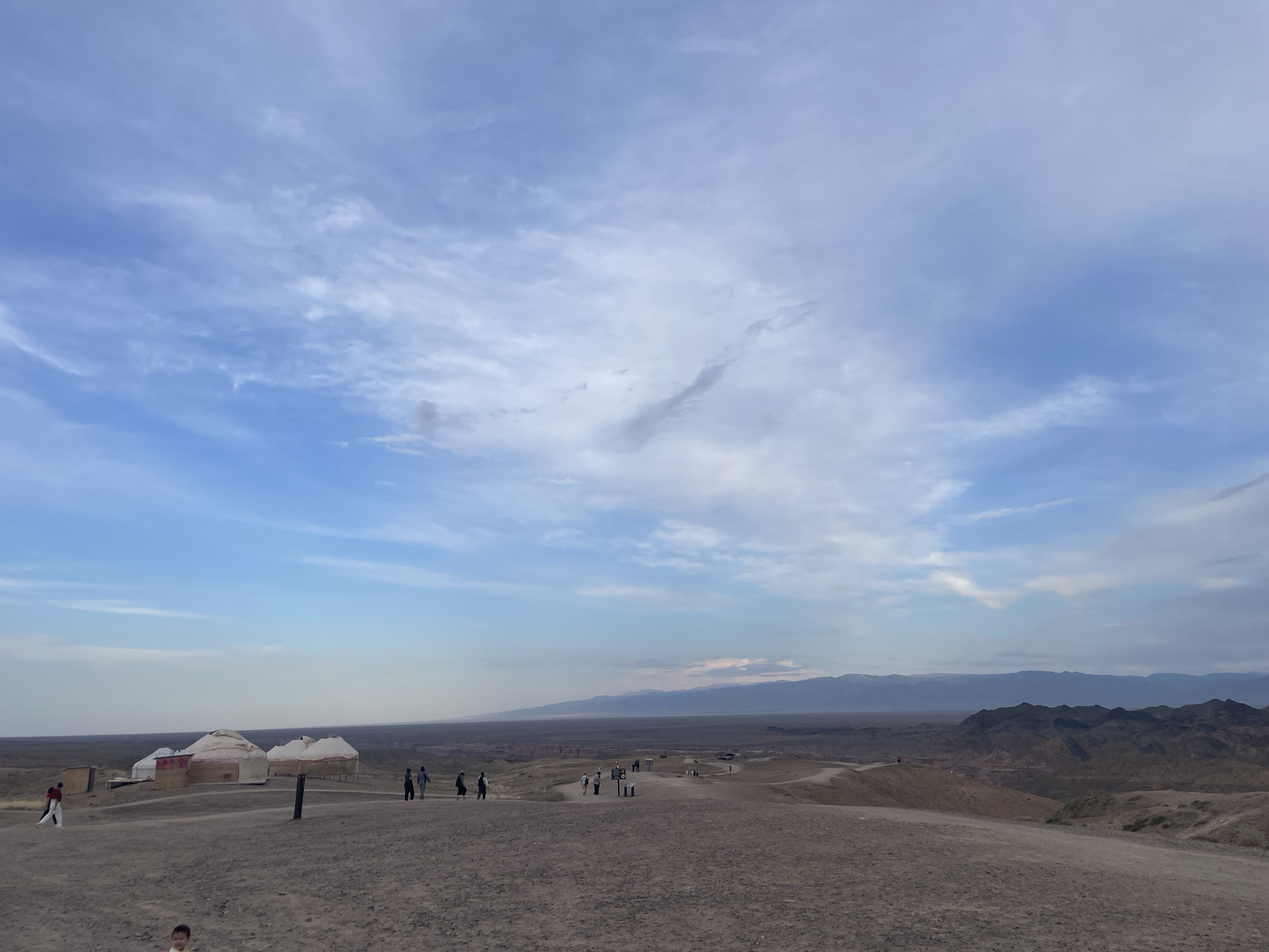 The image depicts a landscape scene in Tamerlik, Kazakhstan. In the foreground, a dusty, barren terrain with scattered vegetation is visible, with a few individuals walking. Several yurts, traditional nomadic dwellings, are situated to the left, suggesting a settlement or tourist area. In the mid-ground, more people are seen strolling along paths that wind through the landscape. The background is dominated by a vast, desolate plain that stretches towards a range of mountains on the horizon, their peaks appearing hazy under a partly cloudy sky. The overall impression is one of a remote, natural environment with signs of human presence.