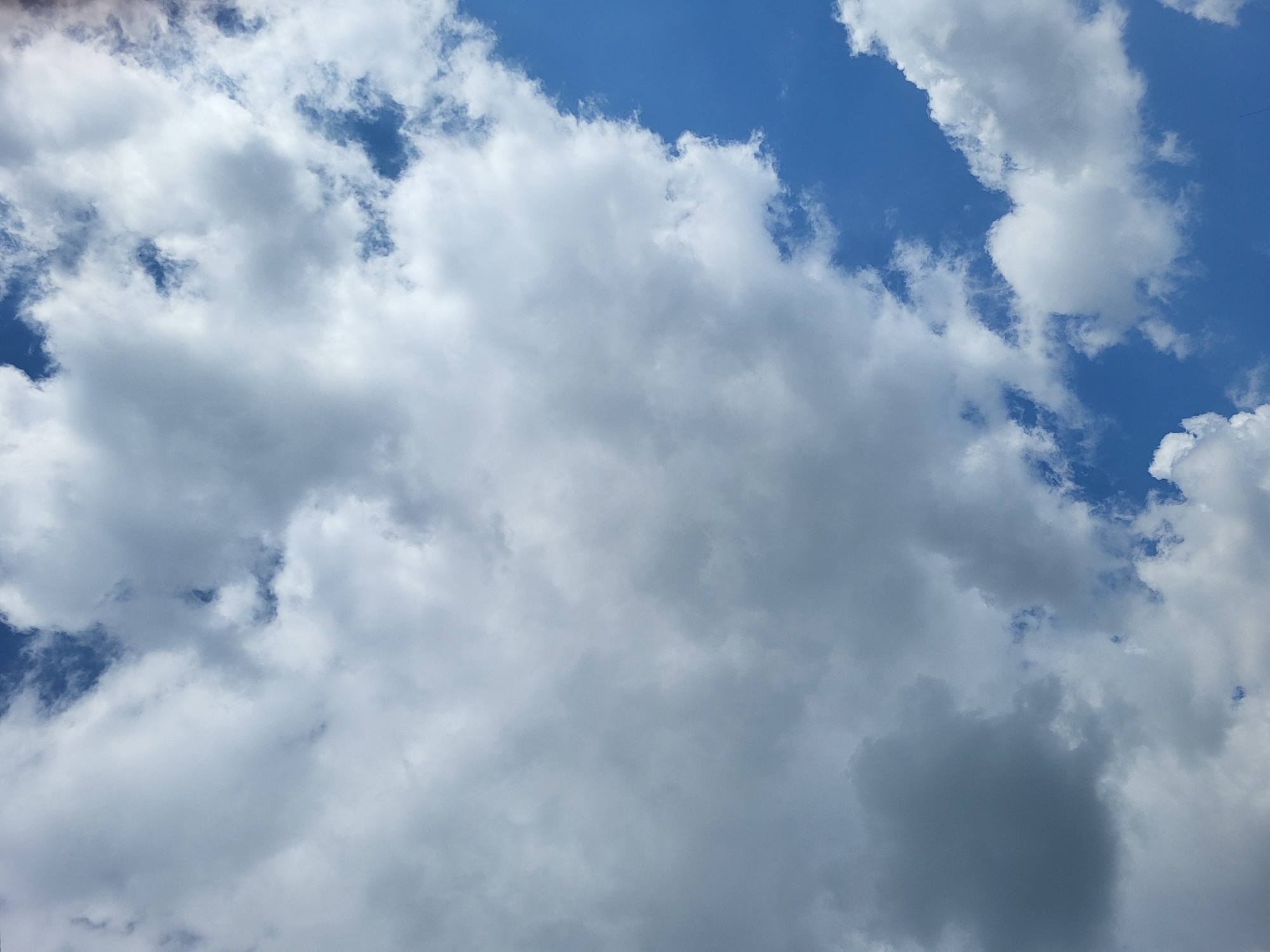 The image shows a daytime sky with a mix of puffy white clouds and patches of blue sky. The clouds vary in size and density, with some appearing thin and translucent, while others are denser and darker. The overall impression is of a partly cloudy day. Location: Unknown, Unknown.