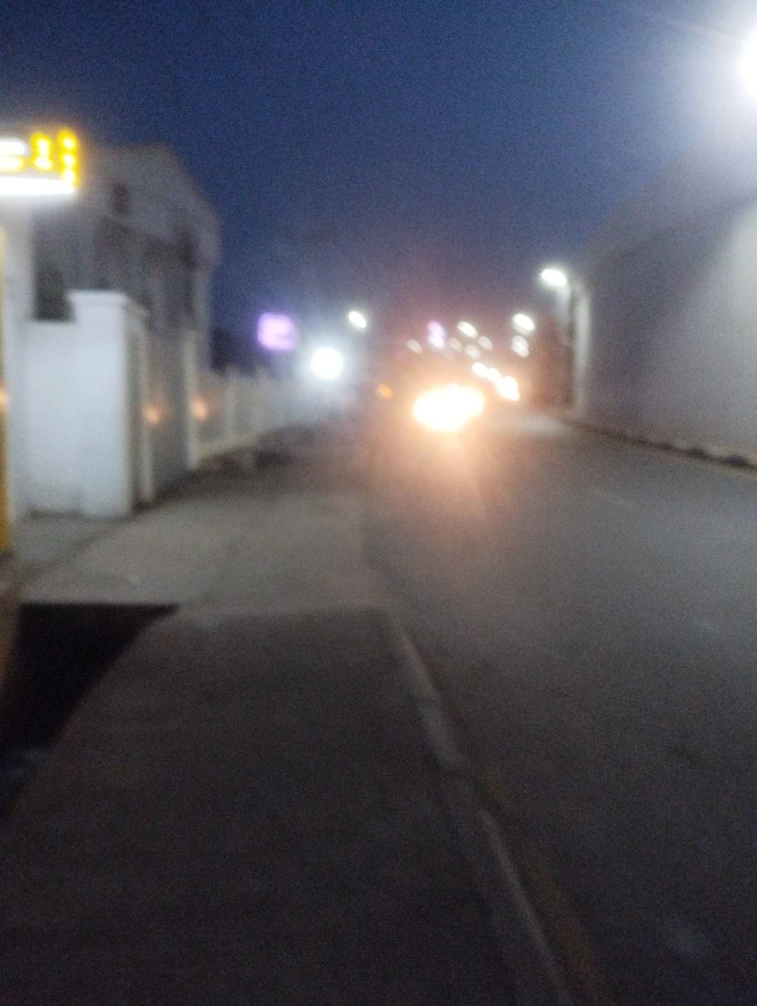 The image depicts a street scene at dusk or night in Jos, Nigeria. The foreground is dominated by a dark, paved road with a slight incline, suggesting a downhill or slight gradient. To the left, a sidewalk area is visible, with a dark, rectangular shadow that could be an open drain or a recessed area. Beyond the sidewalk, a low white wall and a building with yellow, illuminated signage are partially visible. The building appears to be a commercial establishment, possibly a shop or business, with the glowing yellow lights of its sign being a prominent feature.
Further down the street, the scene becomes more blurred due to the low light conditions and possibly motion blur. Streetlights illuminate the road, creating bokeh effects with their bright, circular glows. The headlights of an approaching vehicle are clearly visible, casting a strong, warm light on the road ahead. This suggests that the photo was taken from a stationary or moving perspective, possibly from a vehicle or while walking.
In the background, more lights from other vehicles and possibly buildings can be seen, contributing to the overall ambiance of a moderately busy urban street at night. The sky is a dark, deep blue, typical of twilight or nighttime. The overall mood of the image is somewhat atmospheric and urban, capturing the essence of a street at night with artificial light sources providing illumination. There are no clearly discernible people in the image, but the presence of vehicles indicates ongoing activity. The setting appears to be an urban environment with commercial and residential elements.