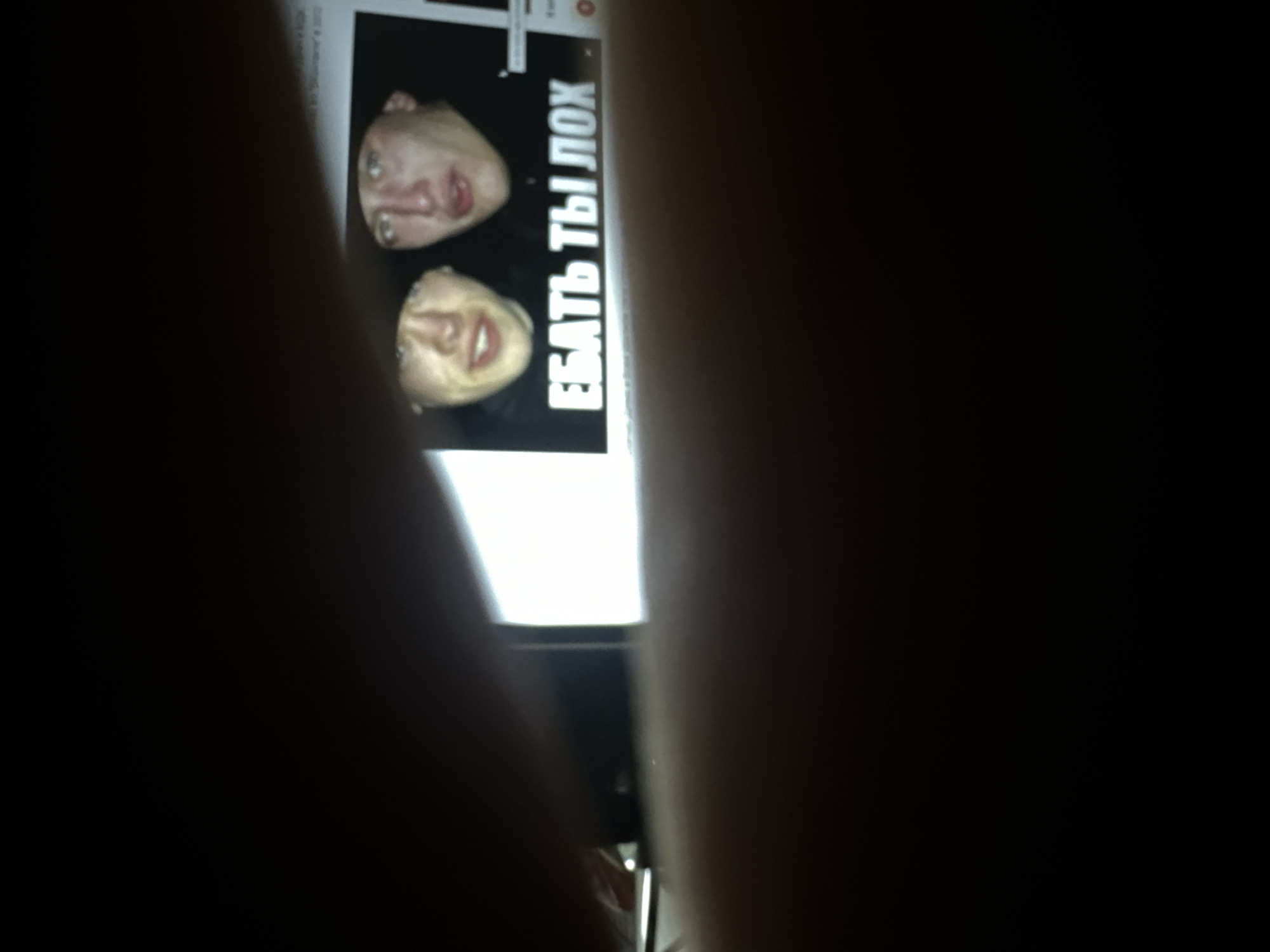 This is a photograph of a computer screen displaying a meme. The meme features two close-up images of people's faces, positioned one above the other. Both faces appear to be looking slightly upwards, with mouths open. To the right of the faces is large, white text on a black background. The text is in Cyrillic and reads "ЕБАТЬ ТЫ ЛОХ". The photograph itself is taken at an angle, with parts of the screen obscured by dark, blurry areas that appear to be foreground elements, possibly hands or other objects. The lighting suggests the image was taken in a dimly lit environment, with the computer screen being the primary light source. There are no visible cues about the time of day, weather, or a specific location, other than the presence of a digital display. The overall impression is that of someone capturing a screenshot or a photograph of a meme they are viewing on a screen.