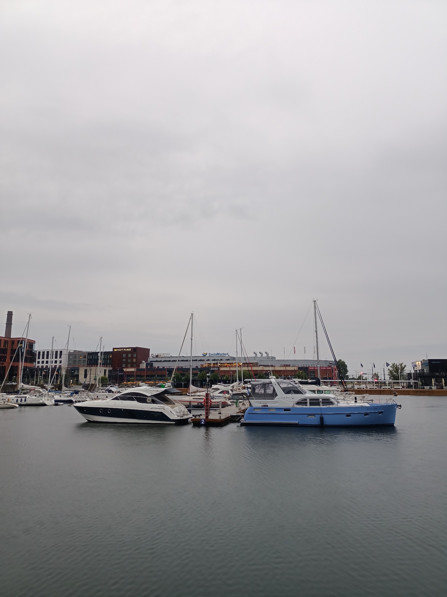 This is a full-shot, eye-level photograph taken on an overcast day of a marina in Tallinn, Estonia. In the foreground, the dark, rippled water of the marina stretches across the frame. Several yachts are docked in the middle ground. On the left, a sleek white and black yacht with a rounded hull is moored. To its right, a larger, two-story blue yacht with a white upper deck is docked. Other yachts and sailboats, some with their masts visible, are also present in the marina. In the background, the city skyline is visible, featuring a mix of modern and industrial buildings. Notable structures include a brick building with a smokestack on the far left, a modern hotel with "HYATT PLACE" signage, and a long building with "SadaMarket" visible on its facade. The sky is a uniform, pale gray. No people are visible in the scene.