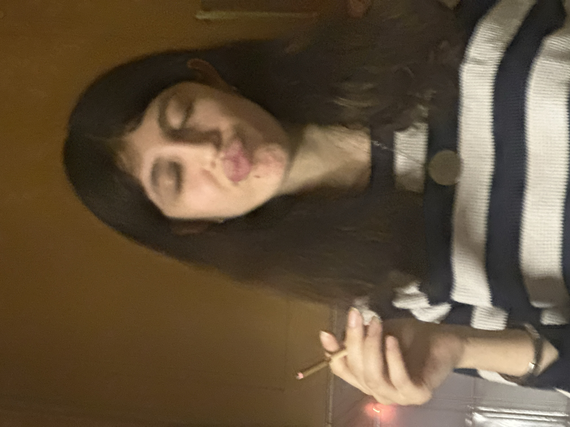 A woman with dark hair is holding a lit cigarette. She is wearing a striped shirt. The background is a plain wall. The location is not discernible from the image.