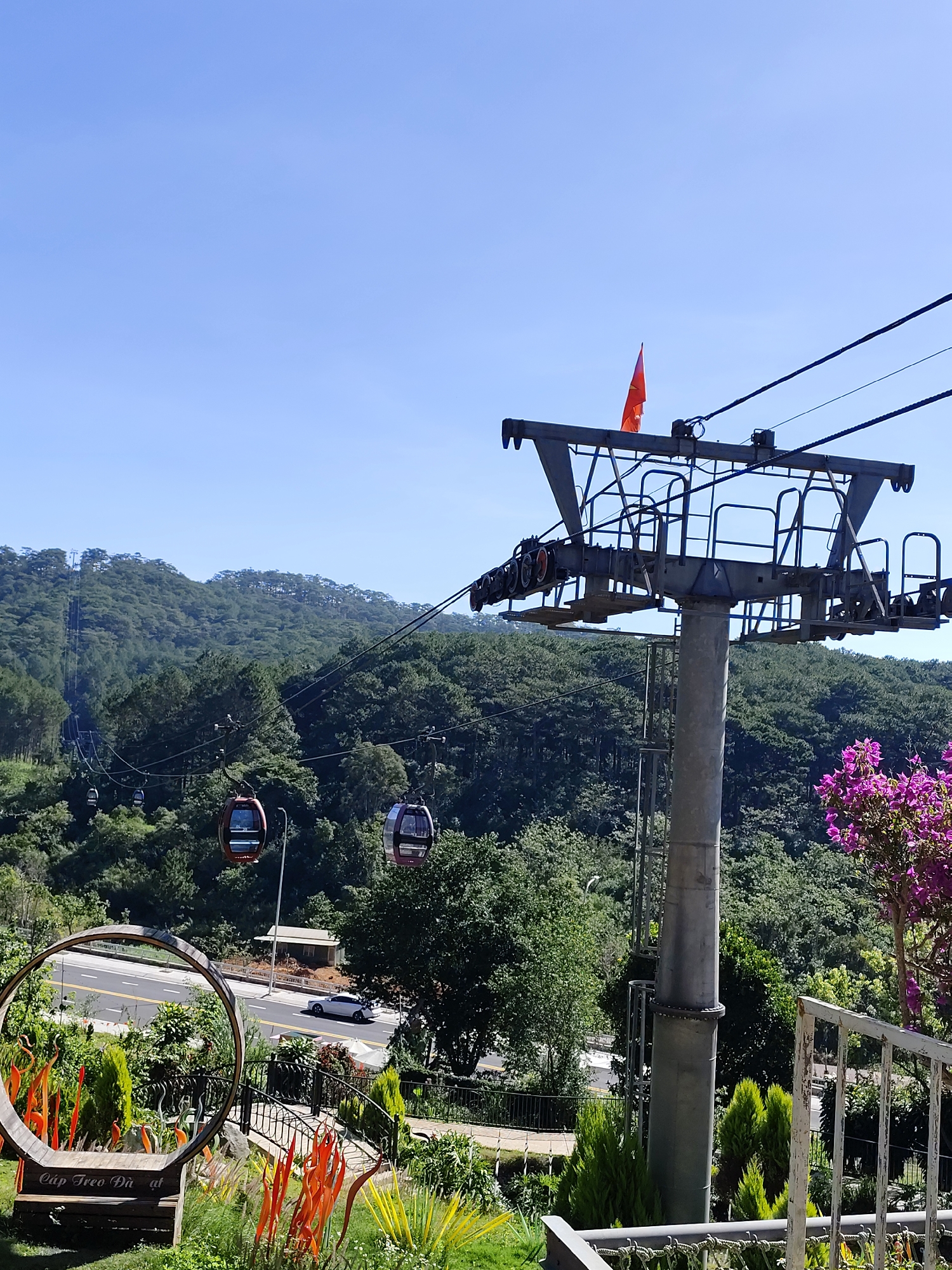 The image shows a cable car system in Da Lat, Vietnam. The support tower of the cable car, topped with a small orange flag, stands prominently in the right foreground. Cable cars are visible moving along the line, ascending a forested hillside in the background. A road with a white car can be seen in the middle ground, passing through a lush garden adorned with orange glass art installations and a decorative wooden circle frame. The sky is clear and blue.
