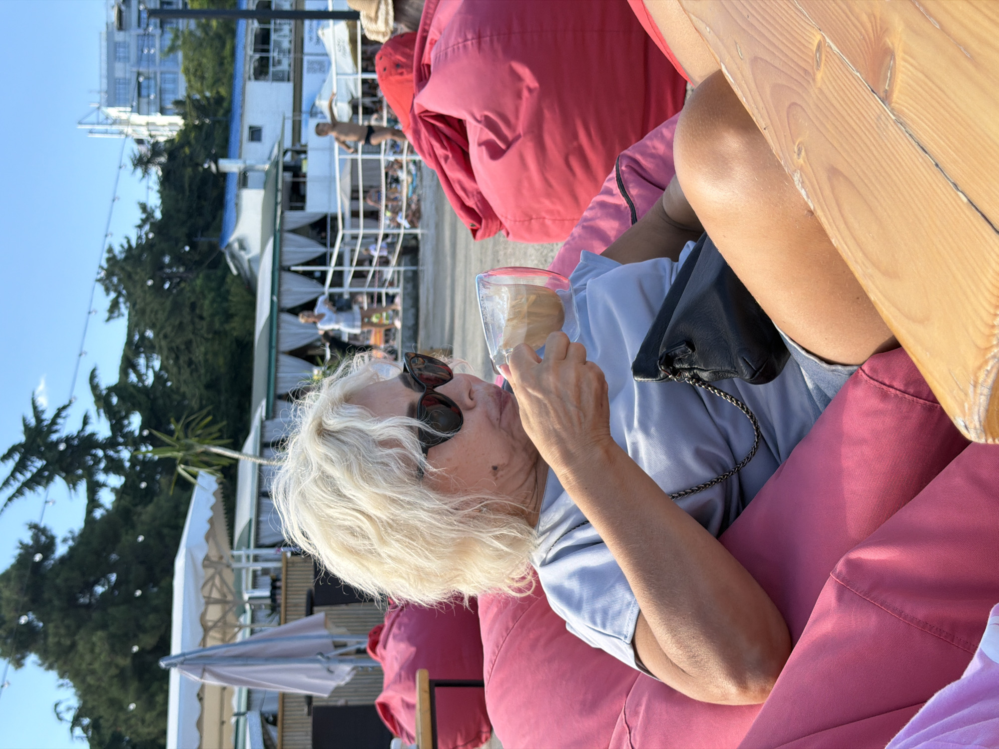 A woman with blonde hair and sunglasses is sitting on a pink beanbag chair, drinking from a glass cup filled with a light-colored beverage. She is wearing a light blue t-shirt and appears to be outdoors, possibly at a beach or outdoor cafe. In the background, there are trees, buildings, and other people, suggesting a public space. A wooden table is visible in the foreground. The scene is lit by natural sunlight, casting shadows and creating a warm atmosphere.