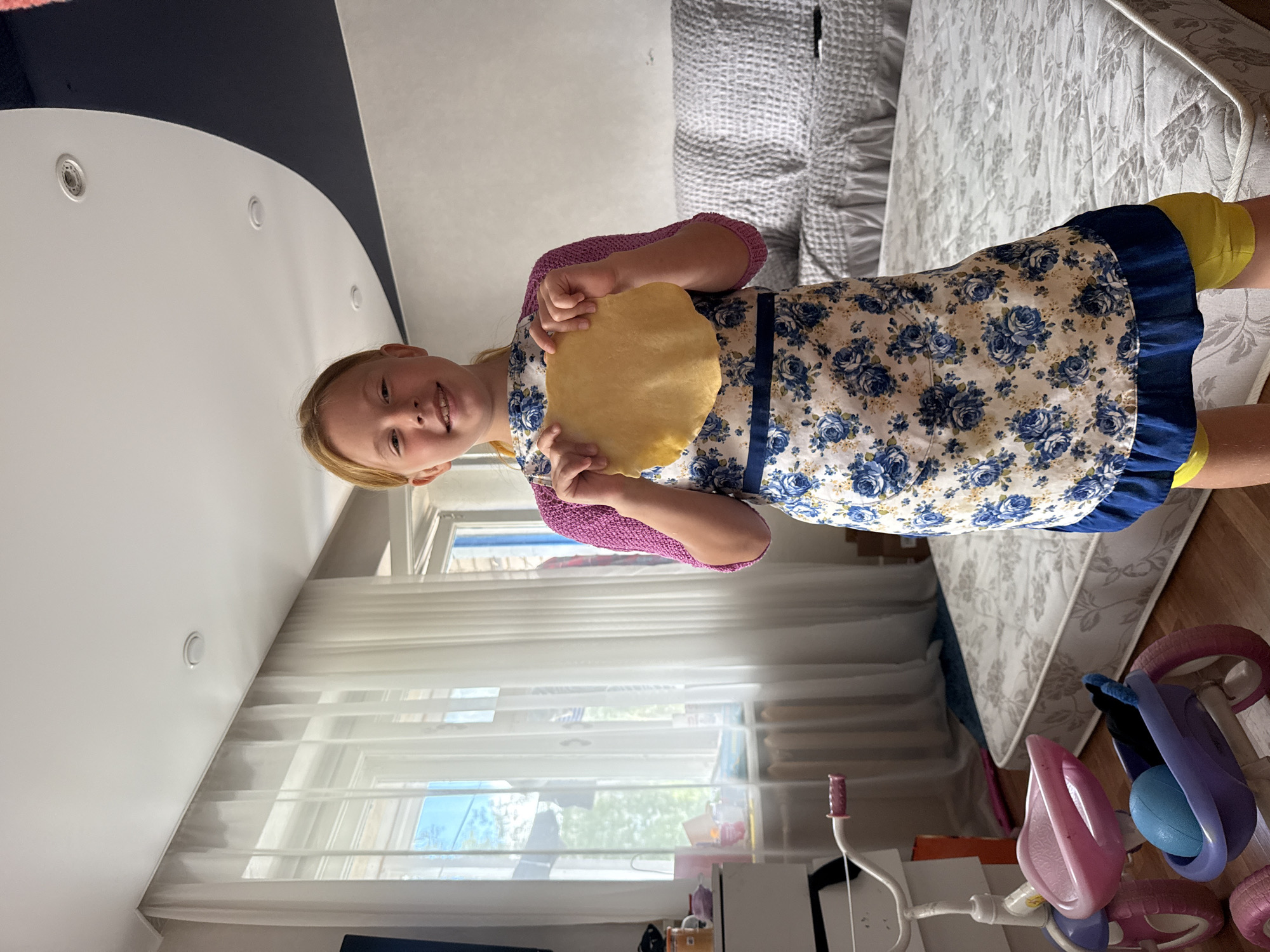 A young girl with blonde hair and fair skin, wearing a floral apron and a pink knitted sweater, stands in a room and holds a round, flat piece of dough. She is smiling directly at the camera. Behind her, sheer white curtains cover a large window. To the left, a white dresser and a child's tricycle are visible. In the background, a gray couch and a mattress lie on the floor. The room has a high ceiling with recessed lighting. The setting is identified as a residential interior in the city of Valday, Russia.