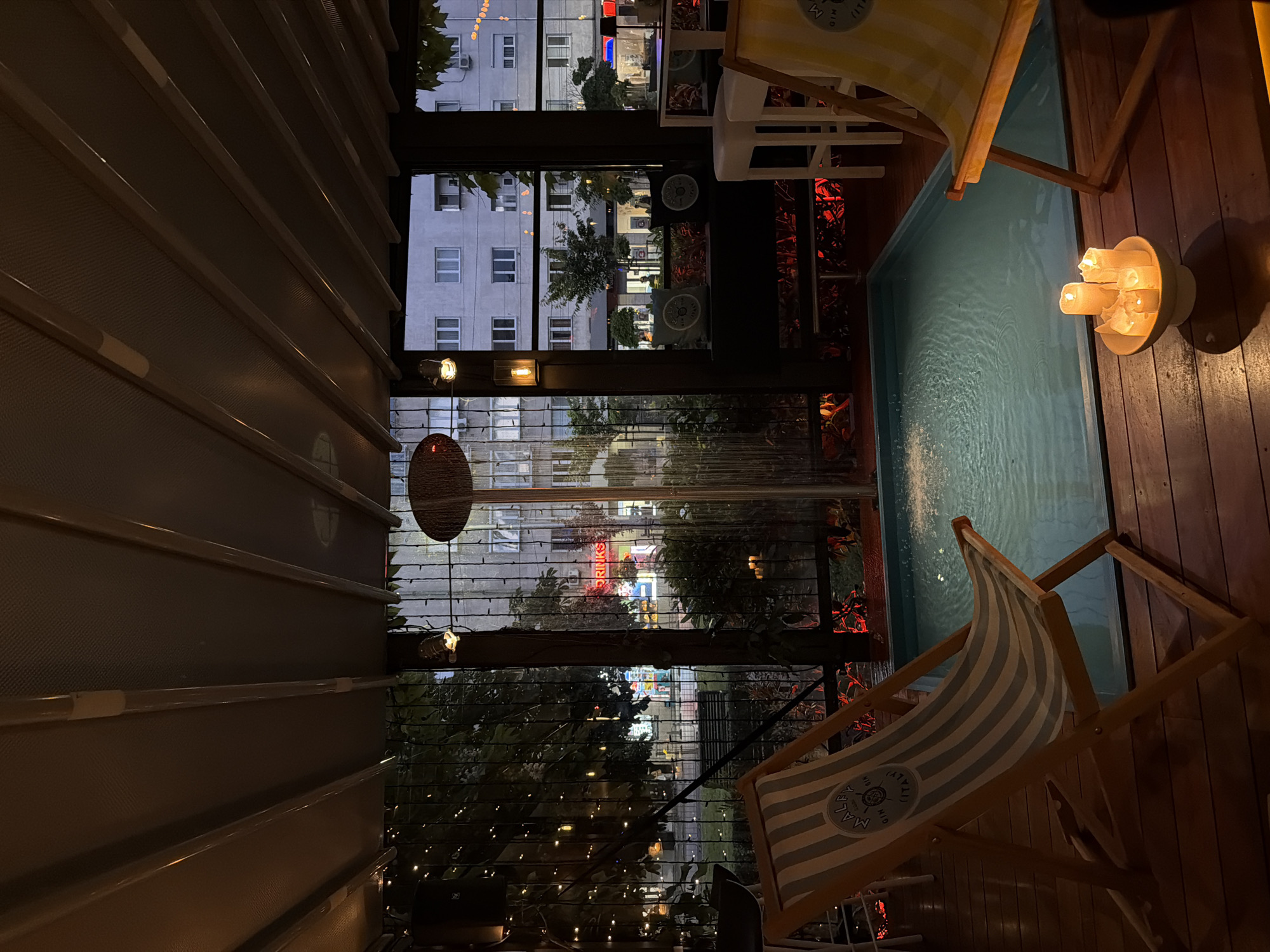 The image shows an indoor space with a view to the outside in the city of Sofia, Bulgaria. The focus is on an outdoor area that appears to be part of a bar or restaurant, with a small, rectangular pool of water to the right. Two beach chairs, one yellow and white striped and the other grey and white striped, are positioned next to the pool. A cluster of lit candles sits on the wooden surface near the pool. Through the window, the buildings of Sofia are visible in the background, illuminated at night. A red neon sign is also visible outside, it may be a sign for "DRINKS". There is a venetian blind on the left side of the image.