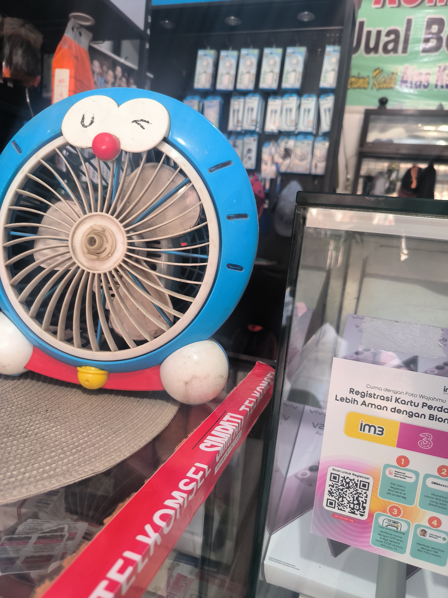 In Tinambung, Indonesia, a Doraemon-themed fan sits on a counter in front of a glass display case filled with cell phone accessories and promotional materials. The fan, primarily blue with white accents and a red nose, is angled to the left. A red and white banner, likely advertising a telecommunications company, is draped diagonally in the foreground, partially obscuring the fan and the display case. The display case contains various items, including what appear to be phone cases and possibly SIM cards, alongside a printed advertisement with a QR code for mobile number registration. In the background, shelves are stocked with packaged electronic accessories, and a sign reads "Jual B," suggesting a retail environment selling goods. The lighting appears to be artificial, illuminating the various products and the fan.