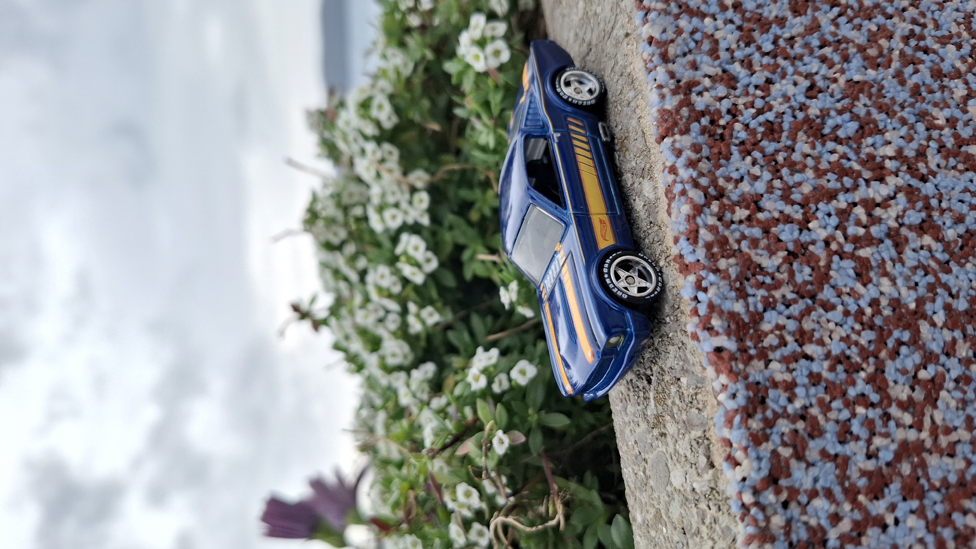 This image depicts a miniature blue Hot Wheels die-cast model, specifically a 1967 Ford Mustang fastback, positioned on a concrete ledge in Boreti, Montenegro. The car features yellow racing stripes on the hood, roof, trunk, and sides, with "Hot Wheels" branding visible on its left flank. It has silver five-spoke wheels with "DREAM+CAR" text on the tires. Behind the car, a lush bed of green foliage with numerous small white flowers is present, with a blurry purple flower visible on the far left. In the background, under an overcast sky, a body of water and a distant landmass are discernible. The immediate foreground, to the right, reveals a wall textured with embedded reddish-brown, light blue, and white aggregate. No people or active interactions are present in the scene.