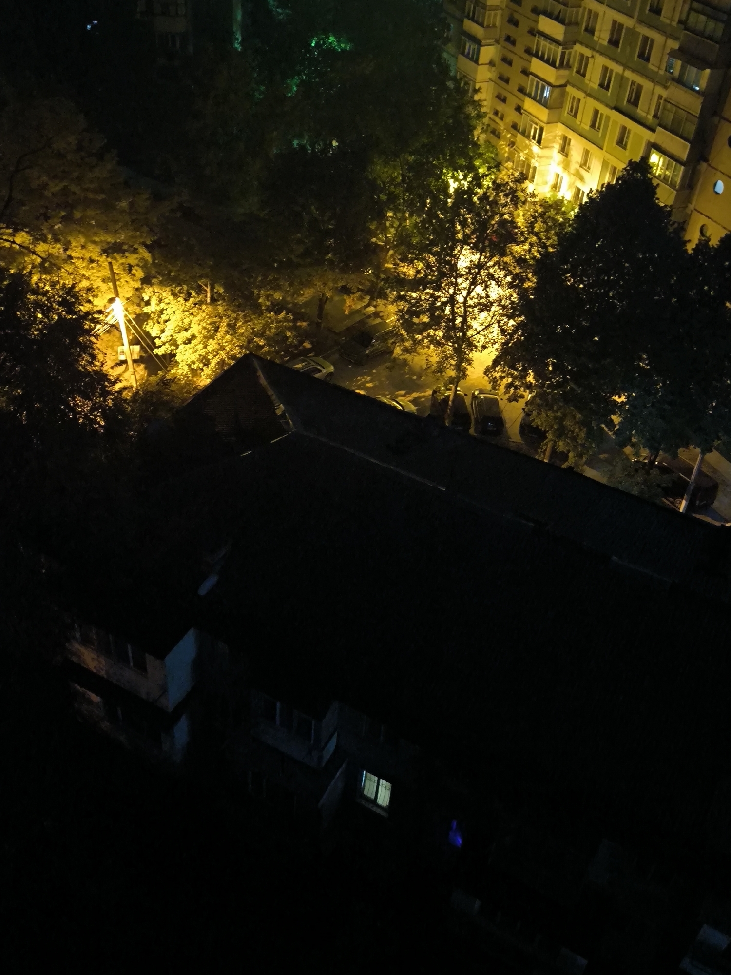This is a nighttime aerial shot looking down on a residential area in Chișinău, Moldova. A large, dark-roofed building occupies the foreground. Behind it, trees are illuminated by streetlights casting a warm yellow glow. Further back, a multi-story apartment building with numerous windows is visible, some lit from within, reflecting the yellow light. Cars are parked along the street between the buildings and trees. The overall scene is dark, with artificial lighting creating pockets of illumination against the night sky.