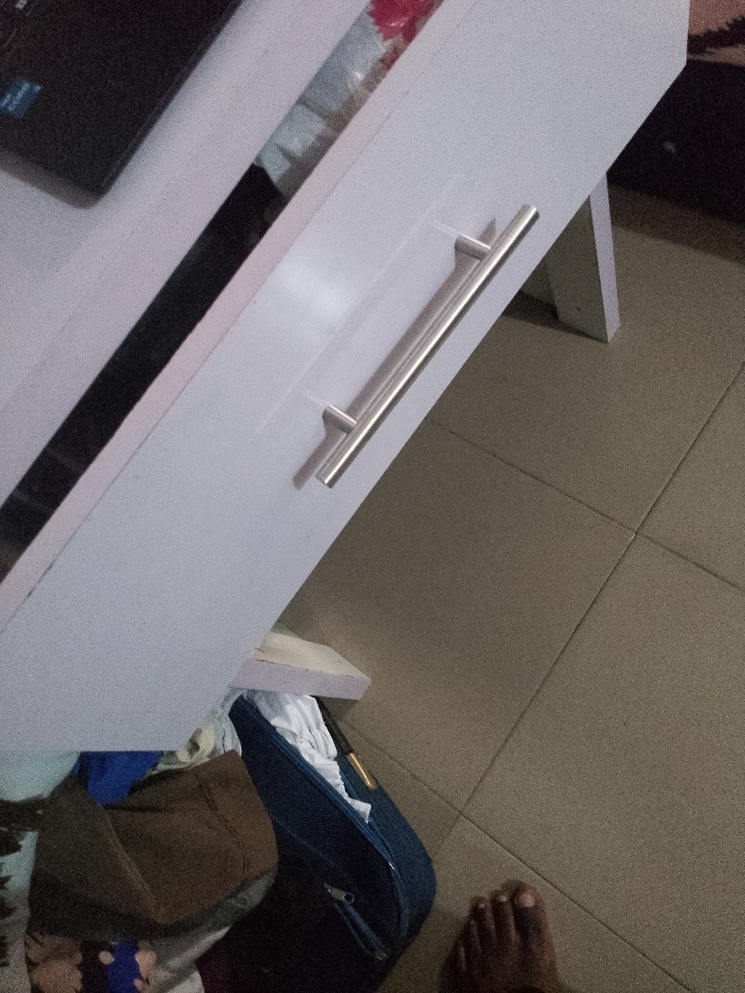 The image shows a close-up of a white cabinet with a silver handle. Below the cabinet, several pieces of clothing are visible, including a brown garment and a blue garment. To the right of the cabinet, a foot is visible on a tiled floor. The background is out of focus but appears to be a domestic setting. The image is taken from a slightly elevated angle.