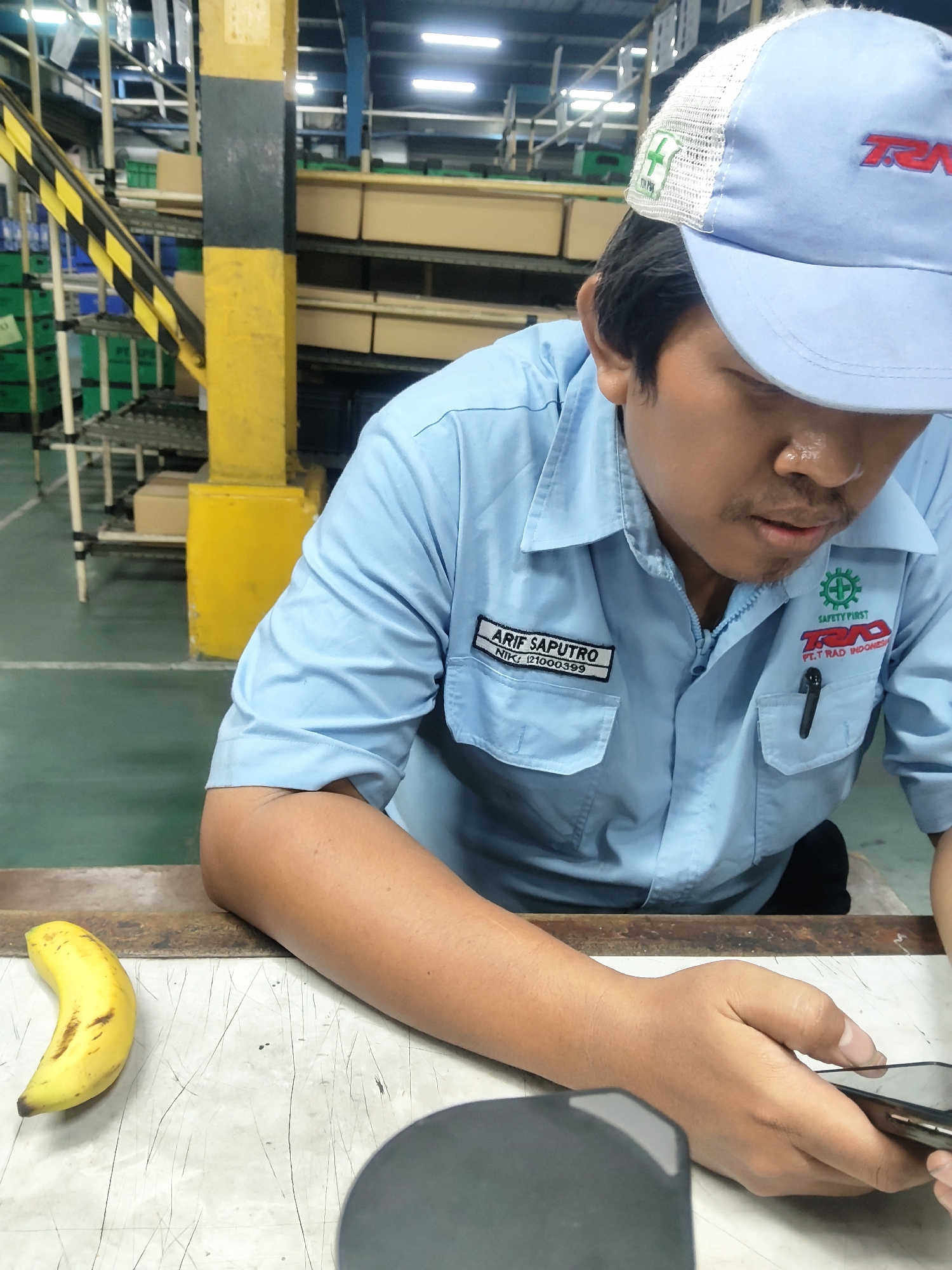 A man wearing a light blue collared shirt and a light blue cap is seated at a workbench in what appears to be a factory setting. The man has dark hair and a mustache. On his shirt, there are two patches: one on the left chest with text "ARIF SAPUTRO NIK: 121000399" and another on the right chest with a green safety logo and text "SAFETY FIRST" and "PT. TRAD INDONESIA". A black pen is tucked into the pocket of his shirt. The man is looking down at a smartphone in his hands. To the left of the man on the workbench, there is a single ripe banana. The workbench itself is light-colored with a textured surface. In the background, there are shelves with boxes and industrial equipment, including yellow and black support beams and a yellow ladder. The overall environment suggests a manufacturing or assembly line facility in Mekarmukti, Indonesia.