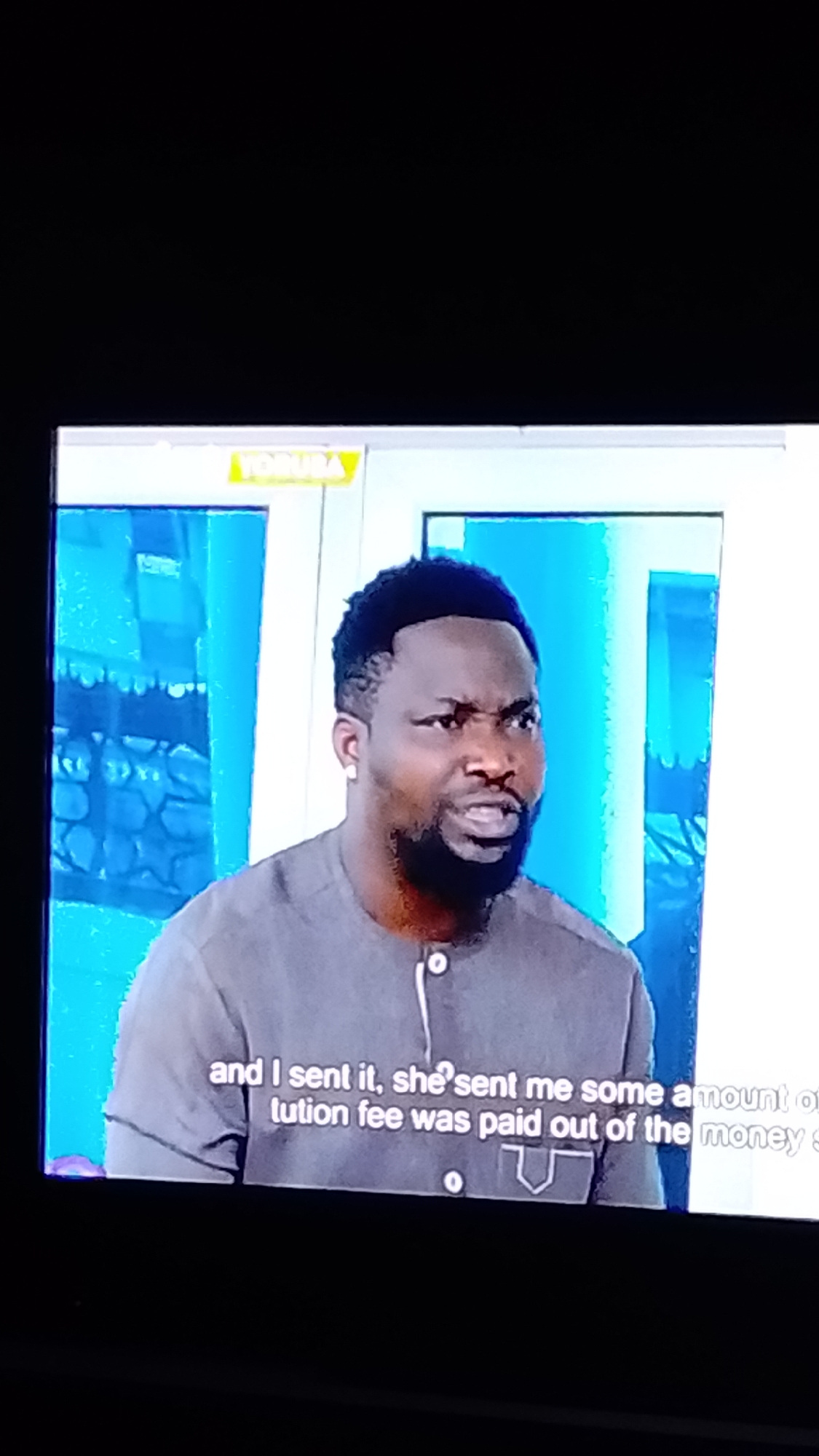 A man with dark skin, a short beard, and a short haircut is looking to the left with a stern expression. He is wearing a grey collared shirt with white stitching and a white button at the neck. The shirt has a decorative pocket on the right side. The background appears to be a television screen with text at the bottom. A yellow logo with the word "YORUBA" is visible in the upper left corner of the screen. The scene appears to be an indoor setting, possibly a studio or a living room. There is no information in the image to suggest a specific location in Lokoja, Nigeria. The man's gaze and expression suggest he is speaking or reacting to something.