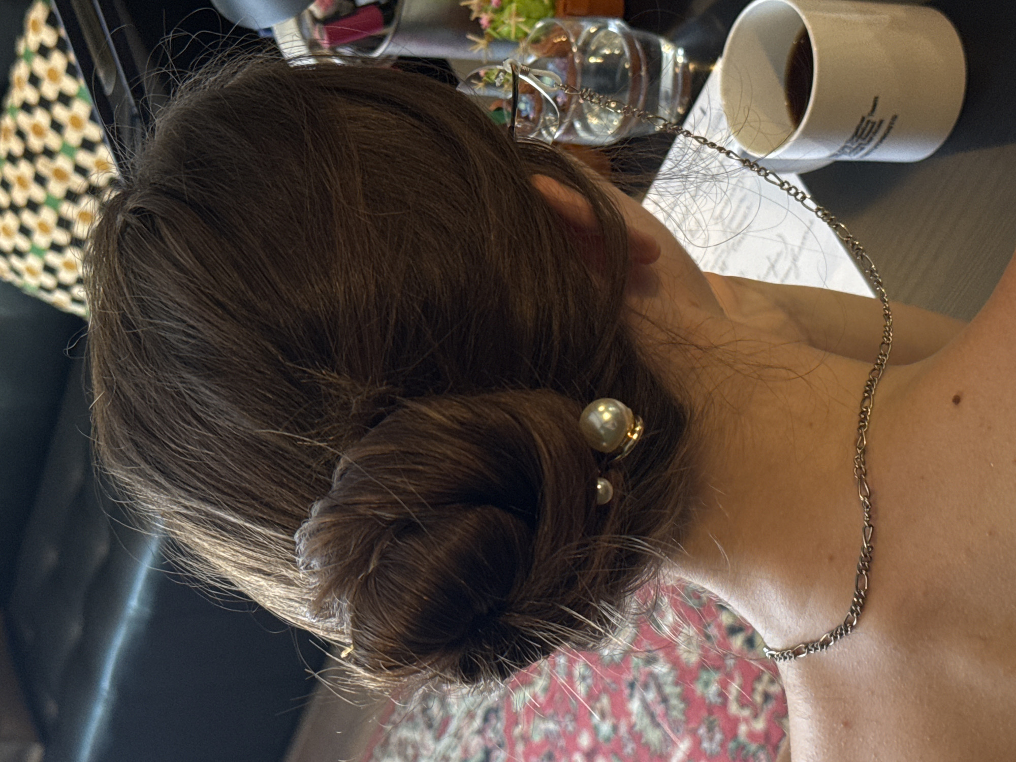 The media file is a close-up, overhead shot of a person's head and neck, with their hair styled in a bun. The person has dark brown hair. Two pearl hairpins are visible in the bun. A delicate silver chain necklace is draped around the person's neck and upper shoulder.
In the background, several objects are visible on a table. To the left, a portion of a black chair and a patterned fabric with yellow and white flowers can be seen. Further back, a clear glass or bottle is partially visible, and to the right, a white mug filled with a dark liquid, likely coffee, is placed on a white surface with some faint text or markings. The lighting suggests it is daytime.
The scene appears to be indoors, possibly in a home or office setting. The overall mood is calm and casual. There is no overt activity or event taking place; the image captures a still moment. The text on the mug is not legible enough to be transcribed. The location context provided is Odesa, Ukraine.