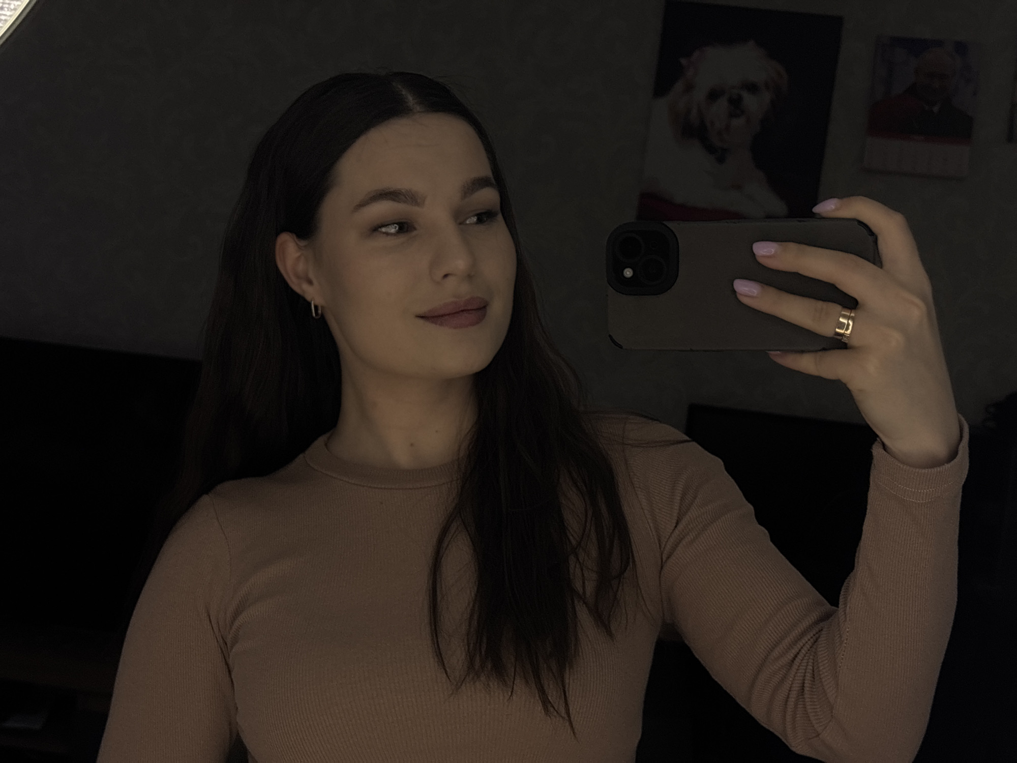 A woman is taking a selfie in an indoor location, possibly a home. The environment is dimly lit. She is wearing a long-sleeved, ribbed brown shirt, gold hoop earrings, and a gold ring. She is holding a smartphone in her right hand, presumably to take the picture. There are pictures on the wall behind her. There are no overt indications of the image's location being in Chaikovsky, Russia.