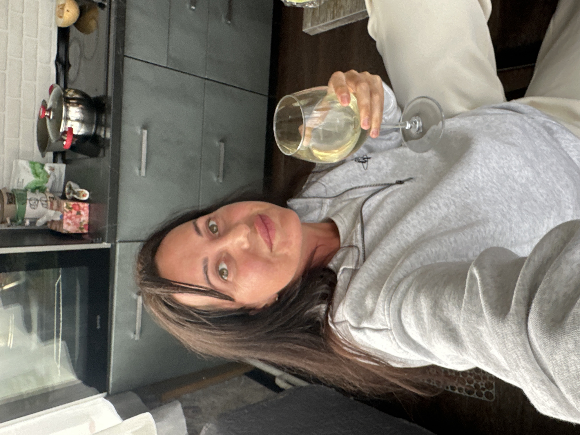 A woman is holding a glass of white wine in her right hand, while sitting on a couch in what appears to be a kitchen. She is wearing a light grey zip-up hoodie and has long brown hair. The kitchen has grey cabinets, a stainless steel pot on the stove, and a brick-tiled backsplash. The scene is brightly lit. The image does not provide any specific information to determine if the location is in Baklashi, Russia.