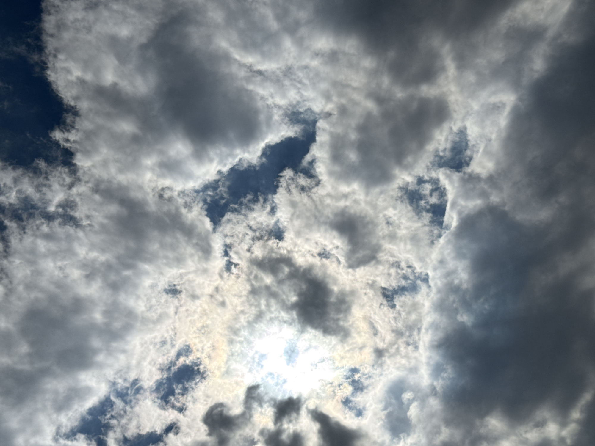 The image displays a sky filled with a mixture of dark and light clouds, with patches of blue visible between them. The sun is partially obscured by the clouds, creating a bright, hazy effect. The scene appears to be an outdoor shot, likely taken from the ground looking upwards, as there is no visible ground or urban landscape. The location is identified as Dzhubga, Russia, though no specific landmarks or features within the city are apparent in this image.