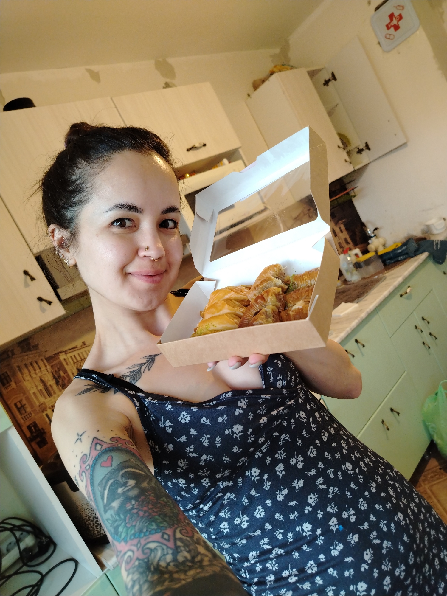 A woman holds a box of baklava in a kitchen. The woman has dark hair pulled back in a bun, fair skin, and several piercings on her ears and nose. She is wearing a navy blue dress with a small white floral print. Tattoos are visible on both arms, including a black floral design on her upper left chest and a more detailed design on her right arm. She is smiling at the camera. The box she holds is cardboard with a transparent window showing the baklava inside. The kitchen has white cabinets along the top and light green cabinets below. There is a depiction of city buildings as a backsplash. A first aid kit is visible on the wall in the background. The city is Noviy, Russia.