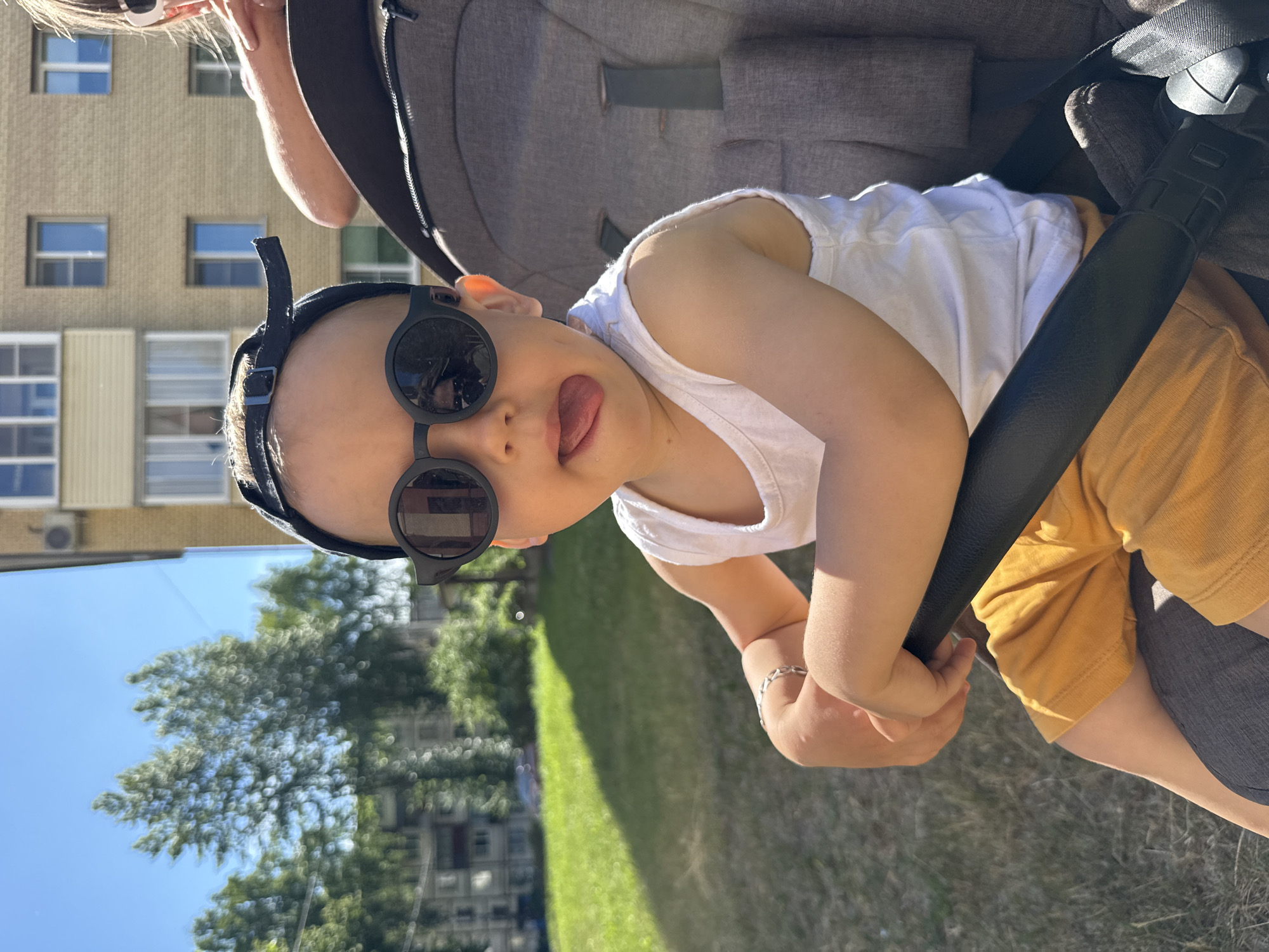 A young child wearing a white tank top, yellow shorts, a black baseball cap, and round sunglasses is seated in a stroller outdoors. The child is sticking their tongue out, and their reflection is visible in the sunglasses. The stroller is brown and has a black handle. In the background, there is a grassy area, trees, and a multi-story building with windows. The sky is clear and blue, indicating a sunny day. The location is identified as Rusanovo, Russia.