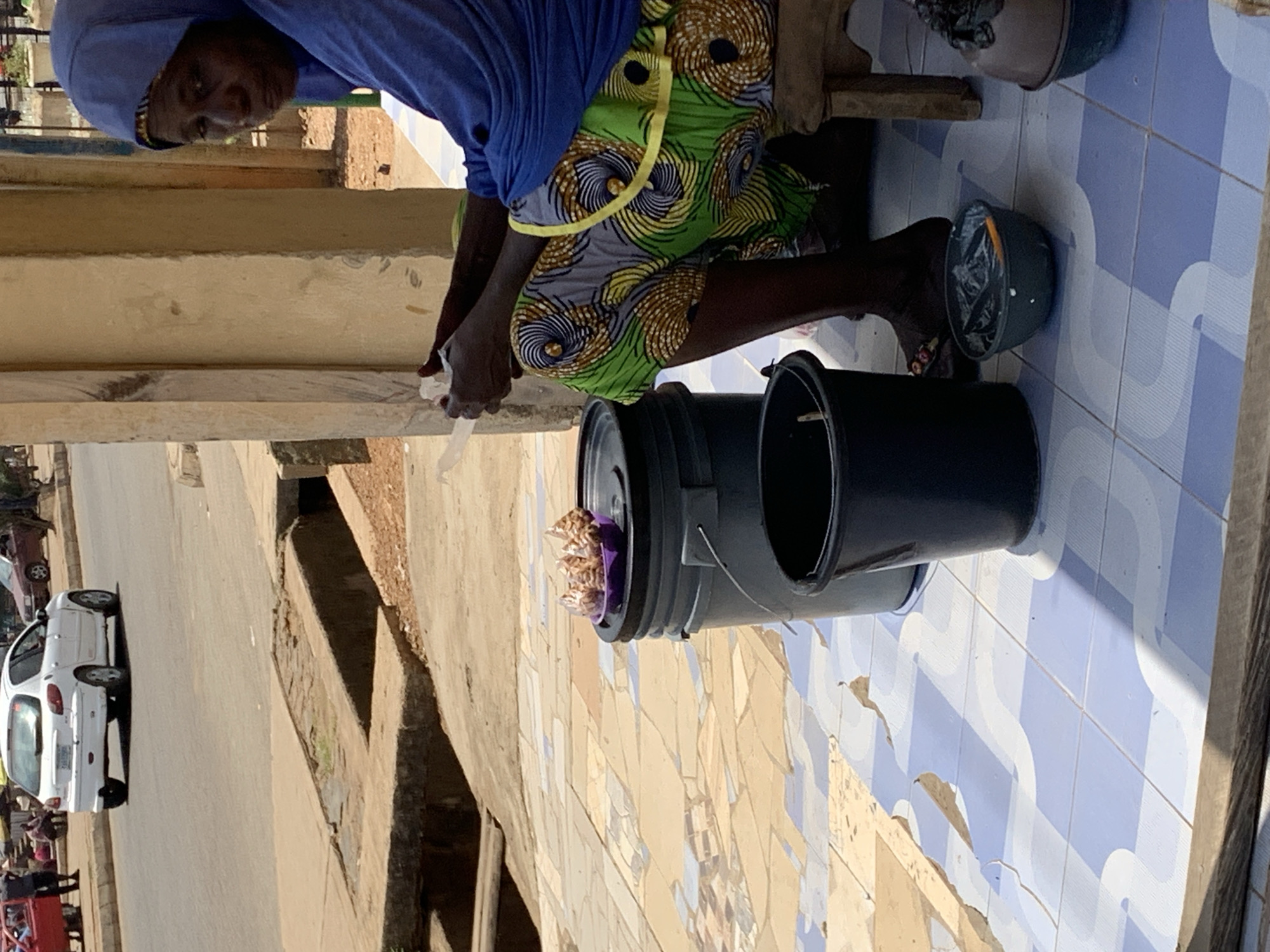 A street vendor sits on a stool outside a building in Rafin Par, Nigeria, packaging food. She is wearing a blue shirt, a patterned skirt, and a headscarf. In front of her are two black plastic buckets. One contains small bags of food. She is packaging food from one container into smaller plastic bags. One filled container and one container on the ground are visible near her feet. A street with cars and pedestrians is in the background. The building and the ground are tiled.
