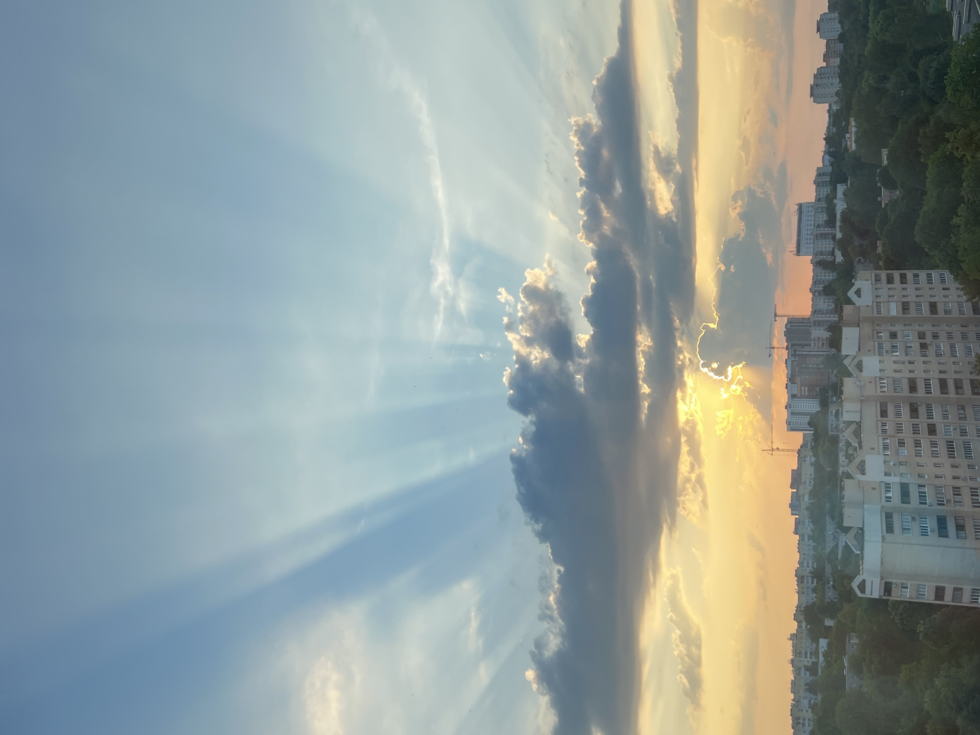 This media file depicts a sky filled with dramatic clouds and crepuscular rays, transitioning into a cityscape as the view moves downwards. The sky is a dominant subject, featuring a clear blue expanse on the left, gradually shifting to warmer tones of yellow and orange near the horizon on the right, indicating either sunrise or sunset. Dark, textured clouds are scattered across the sky, with the sun's rays piercing through them, creating visible beams of light.
Below the sky, a dense urban landscape is visible. Numerous apartment buildings, characteristic of Eastern European architecture, are lined up along a sloping terrain. The buildings are multi-story, with visible windows and balconies, suggesting a residential area. Lush green trees partially obscure the lower portions of the buildings, adding a touch of nature to the urban setting. A construction crane is faintly visible in the distance, hinting at ongoing development.
The overall mood is serene and picturesque, with the dramatic sky providing a sense of awe. The lighting suggests it is either early morning or late evening. The location context of Minsk, Belarus, aligns with the architectural style of the buildings. No visible text is present in the image.
