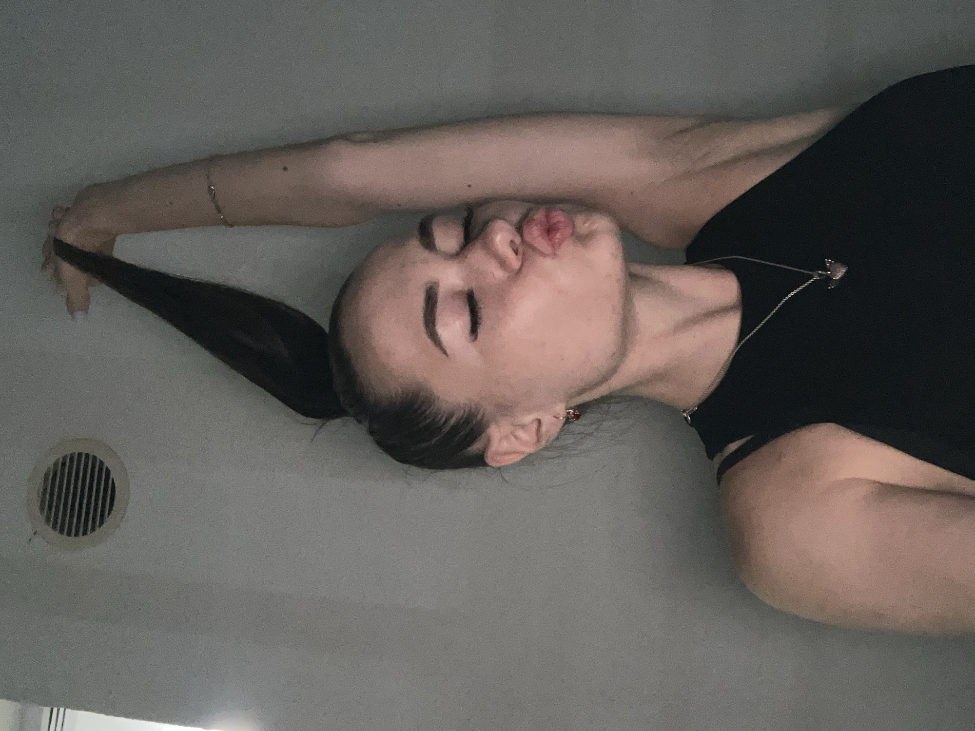 A young woman with her hair pulled up high in a ponytail poses for a selfie, puckering her lips towards the camera. She is wearing a black tank top and a necklace with a heart-shaped pendant. Her right arm is raised above her head, holding her ponytail. The background appears to be a plain, light-colored wall with a circular vent near the top left. The lighting is dim and appears to be artificial. The location is unknown, within the city of Unknown.