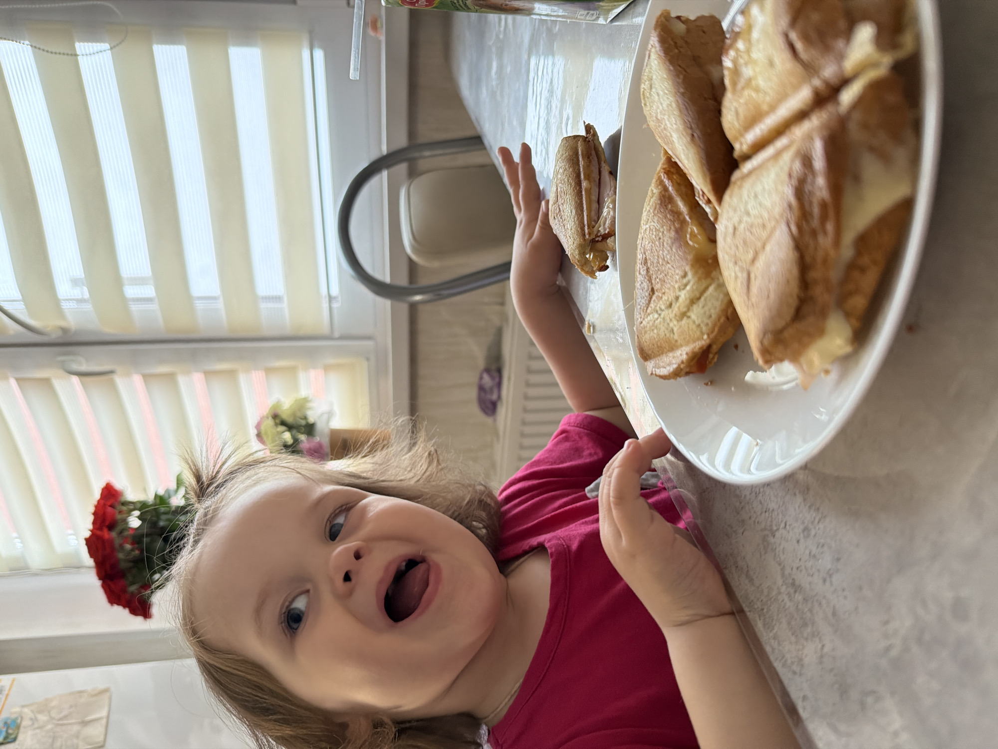 A young child wearing a pink shirt stands next to a table in a bright room. The child has blonde hair with a small ponytail on top of her head and is opening her mouth wide with excitement. On the table next to her is a plate containing several grilled cheese sandwiches. Behind the child, there is a window with white blinds and a bouquet of red flowers on the windowsill. A white chair is partially visible near the table. The scene appears to be indoors, possibly in a kitchen or dining area. This image was taken in the city of Chaikovsky, Russia.