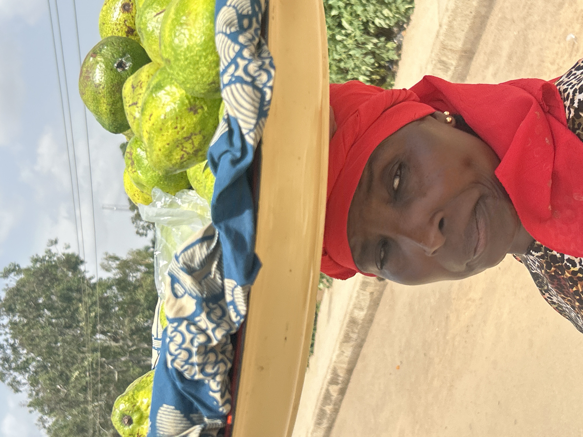 A vendor wearing a red headscarf and animal print shirt stands to the right of a tray overflowing with green avocados and covered in a blue patterned cloth. The vendor looks directly at the camera. The tray appears to be made of a tan or light yellow material and sits between the vendor and the avocados. Some of the avocados are contained in clear plastic wrap. In the background, a blurred street scene with trees and a cloudy sky is visible. This scene is likely in Jos, Nigeria.