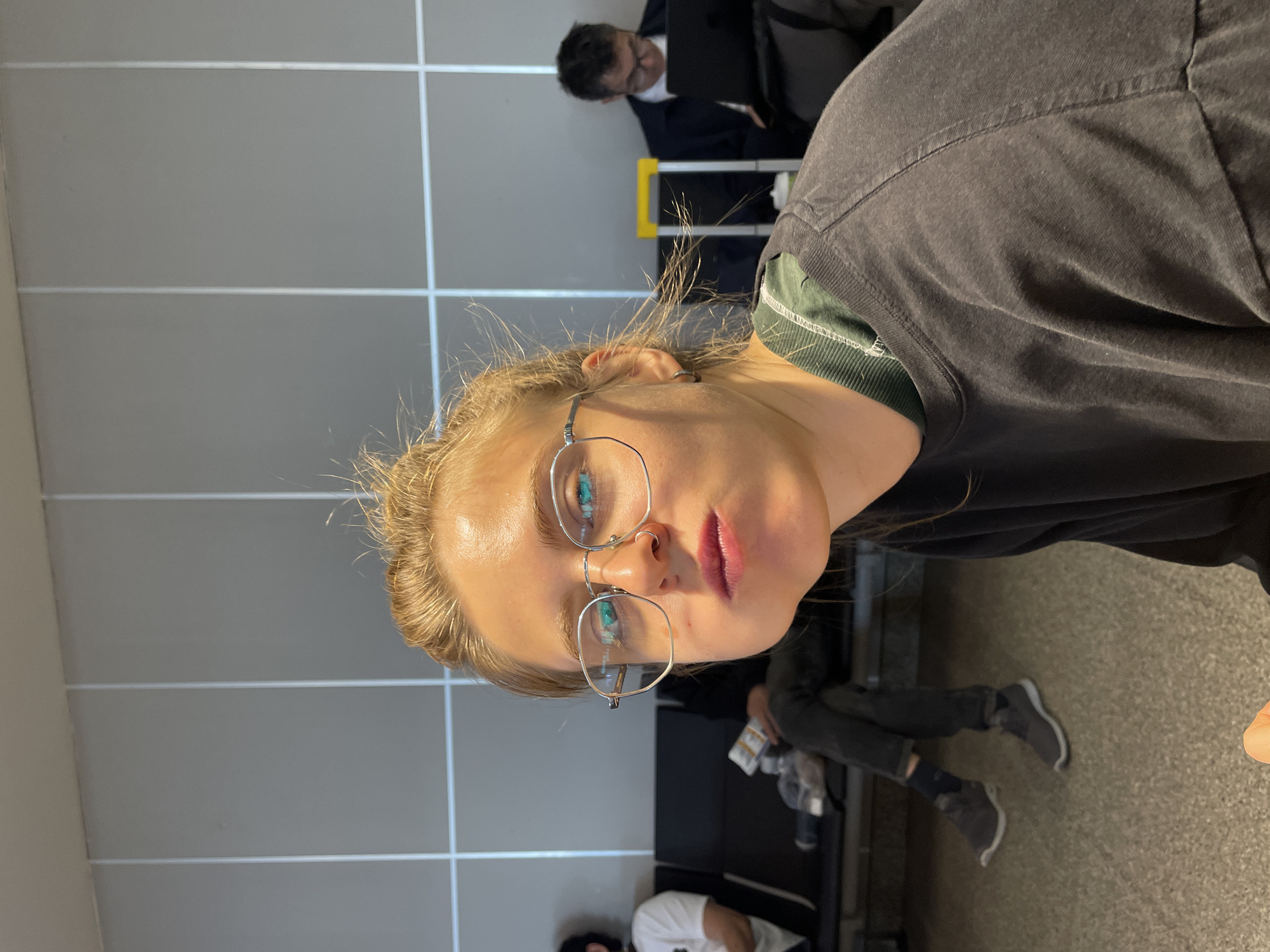 The image presents a close-up view of a woman, seemingly in an airport waiting area. She is wearing glasses and a dark-colored t-shirt over a green, possibly corduroy, top. Her hair is blonde and appears somewhat disheveled.
In the background, other people are visible, seated in rows of chairs typical of airport terminals. One man is wearing a suit. The backdrop consists of a gray tiled wall. Based on the appearance of the waiting area and other people in the picture, it is likely that this photo was taken in a public waiting area. There is not enough information available in the picture to determine that the location is in Tashkent, Uzbekistan.