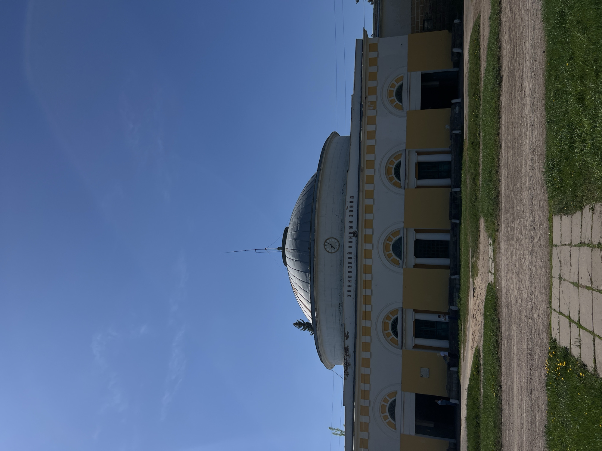 The image shows a building in Sloboda, Russia. The building is two-storied with a rounded dome on top. The building's facade is white with yellow accents around the arched windows. There is a clock visible on the dome. Text is written in Russian on the dome, part of which reads "Khrenovsky Stud Farm." A paved walkway and grass area are visible on the right side of the frame. The sky is blue with some wispy clouds. Several people can be seen standing in front of the building.