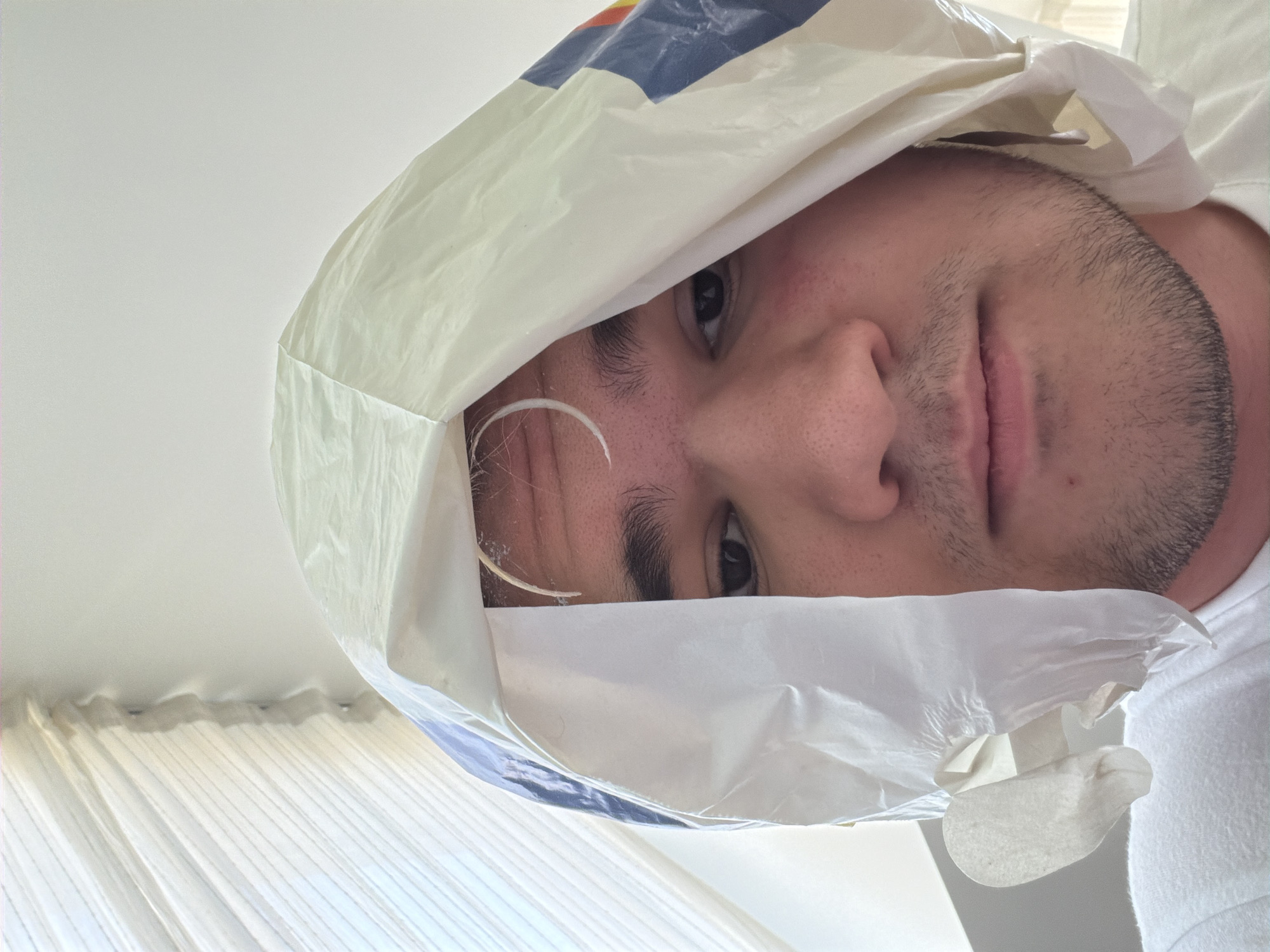 A man is wearing a white plastic shopping bag over his head, obscuring most of his face. Only his eyes, nose, and upper lip are fully visible, along with parts of his forehead and beard. A white object resembling a plastic tie is visible on his forehead, inside the bag. He is wearing a white t-shirt, and white curtains are visible in the background. The scene appears to be indoors. The location cannot be determined from the image.