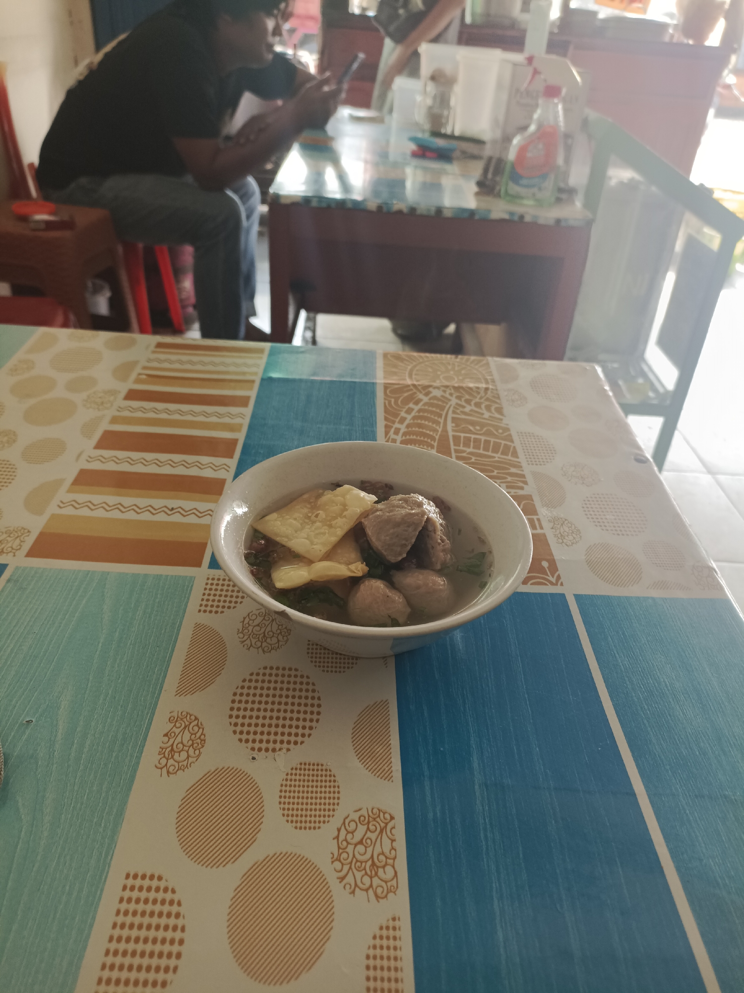 In Sangatta, Indonesia, a bowl of bakso, a popular Indonesian meatball soup, sits on a table covered with a patterned blue and tan tablecloth. The bowl contains several meatballs and crispy fried wontons in a broth, with some green herbs visible. In the background, a person wearing a black t-shirt and jeans is seated, looking at their phone. The environment appears to be an indoor eating area, with other tables and chairs visible. A bottle of hand sanitizer is on a table in the background.