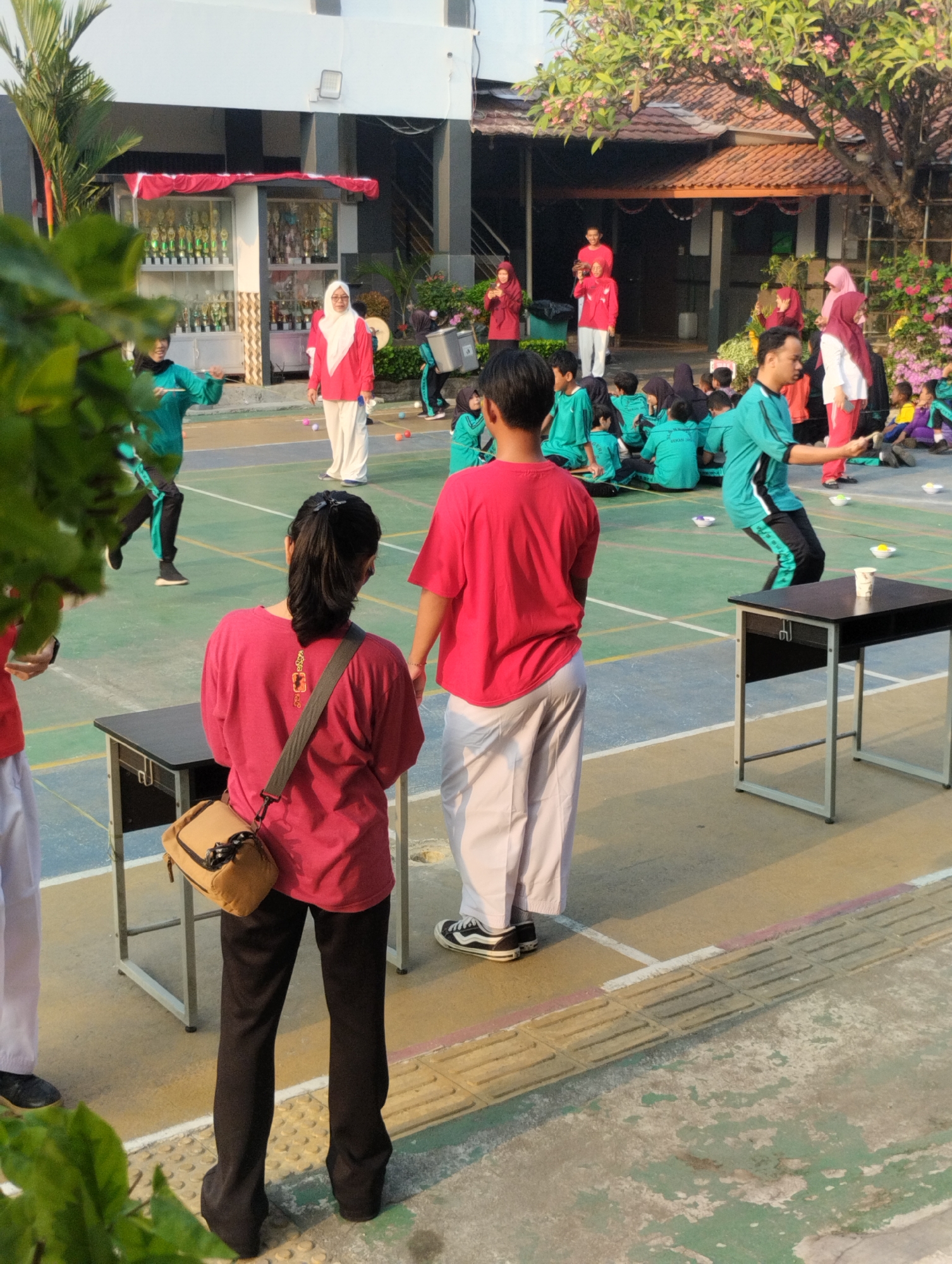This is a medium shot, taken from a low angle, of an outdoor event likely occurring at a school in Bekasi, Indonesia. In the foreground, two individuals stand facing away from the camera, wearing red shirts and white pants. The person on the left has a brown crossbody bag. To their right, a dark table with a white cup sits on it. In the background, several groups of people are visible, many wearing green shirts and headscarves. Some are seated on the ground, while others are standing and participating in an activity that appears to involve throwing or tossing small colored balls. A building with a red tiled roof and trophies displayed in glass cases is visible behind some of the participants. Trees with pink flowers line the right side of the scene. The ground is paved with a green surface marked with white and yellow lines. The lighting suggests it is a sunny day.