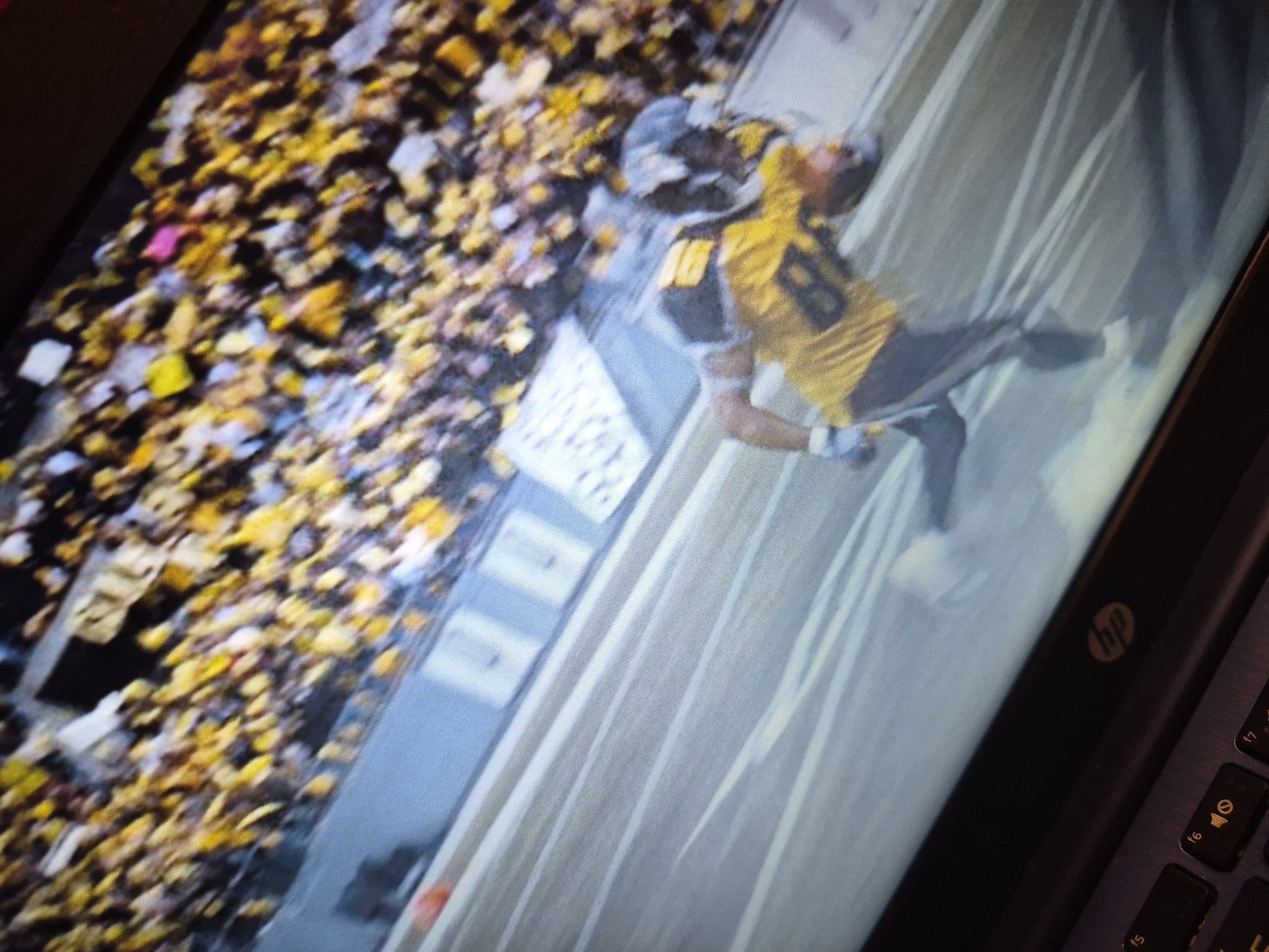 The image is a close-up shot of a laptop screen displaying a sports broadcast. The broadcast shows an American football game, with a player in a yellow jersey with the number 80 on it, wearing a helmet, running on a field. The background consists of a blurred crowd wearing predominantly yellow and black clothing. The stadium appears to be outdoors. The laptop itself is visible in the foreground, with a portion of the keyboard and the HP logo showing. The image is slightly blurry, suggesting it was taken at an angle or without optimal lighting conditions. There are no discernible landmarks or indicators that would place the scene in Sandwell, United Kingdom.