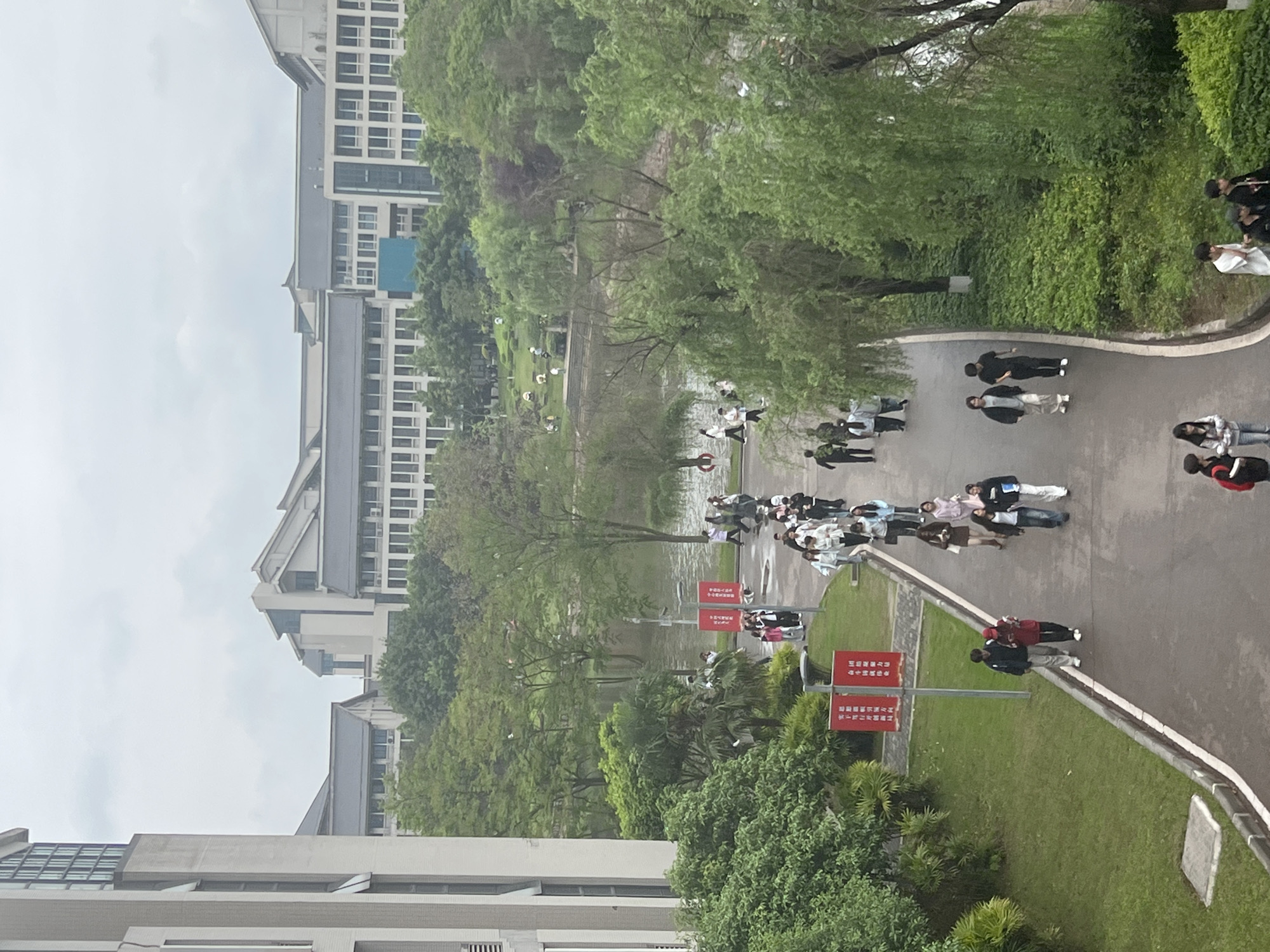 The image shows a view from an elevated position, likely a building, overlooking a section of what appears to be a university campus in Jiangjin, China. Several buildings with light-colored facades and multiple windows are visible in the background and foreground. A body of water, possibly a lake or wide river, runs through the center of the scene. Trees and other greenery line the banks of the water and are interspersed throughout the campus. A paved pathway runs along the side of the water, and groups of people are walking along it. Several red signs with Chinese characters are near the pathway. The sky is overcast.