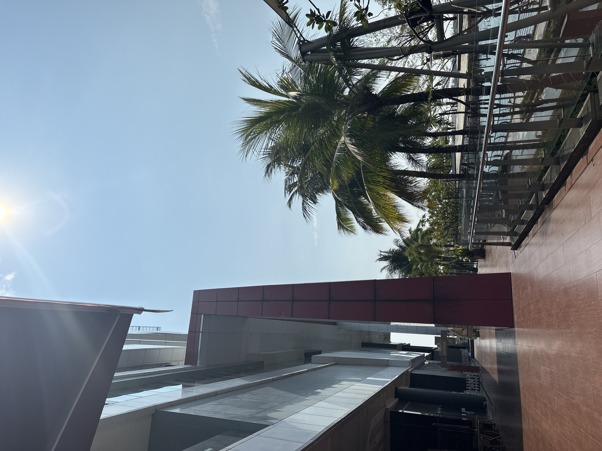 The image depicts an urban landscape in North Jakarta, Indonesia, likely near a modern building complex. The perspective is from a low angle, looking upwards toward a clear blue sky and the sun, which is causing lens flare. Several palm trees are visible, both close and further away. A modern building is in the foreground, featuring a red-paneled section and white, tile-like surfaces, possibly a facade or a roof. A walkway or a balcony with metal railings and glass panels is adjacent to the trees. There are hints of activity below, but people are obscured.