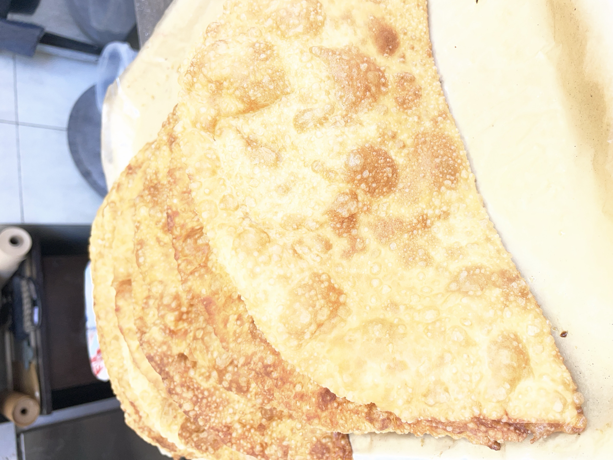 A close-up, high-angle shot shows a stack of golden-brown, deep-fried pastries. The pastries have a flaky, bubbly texture and are arranged in a curved line, with the front pastry occupying the majority of the frame. In the blurred background, a counter and some rolls of paper are visible, suggesting a commercial kitchen or food service setting. There are no people or explicit activities depicted. The setting is not identifiable as Sochi, Russia.