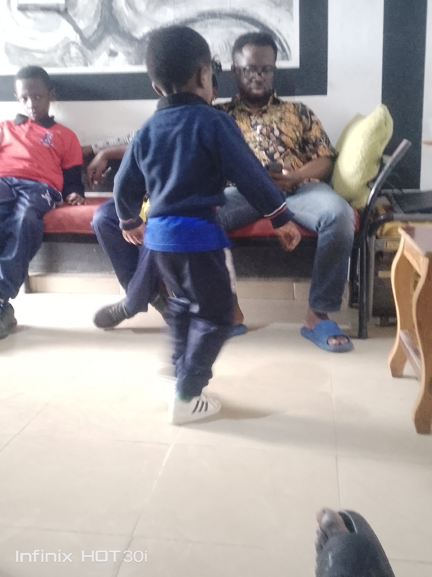 The image shows a group of people in an indoor setting. In the foreground, a young child is walking away from the camera, wearing a dark blue sweater and pants with a blue band around the waist. They have white sneakers with three black stripes on them. To the left, another child is sitting on a red couch, wearing a red shirt with dark sleeves and dark pants. Behind them, a man is sitting on the same couch, wearing glasses and a patterned shirt, holding a phone. A yellow cushion is visible behind him. The floor is tiled. The scene appears to be casual and relaxed, possibly a home or a waiting area. There are no obvious indicators of the time of day or weather. The text "Infinix HOT 30i" is visible at the bottom left of the image.