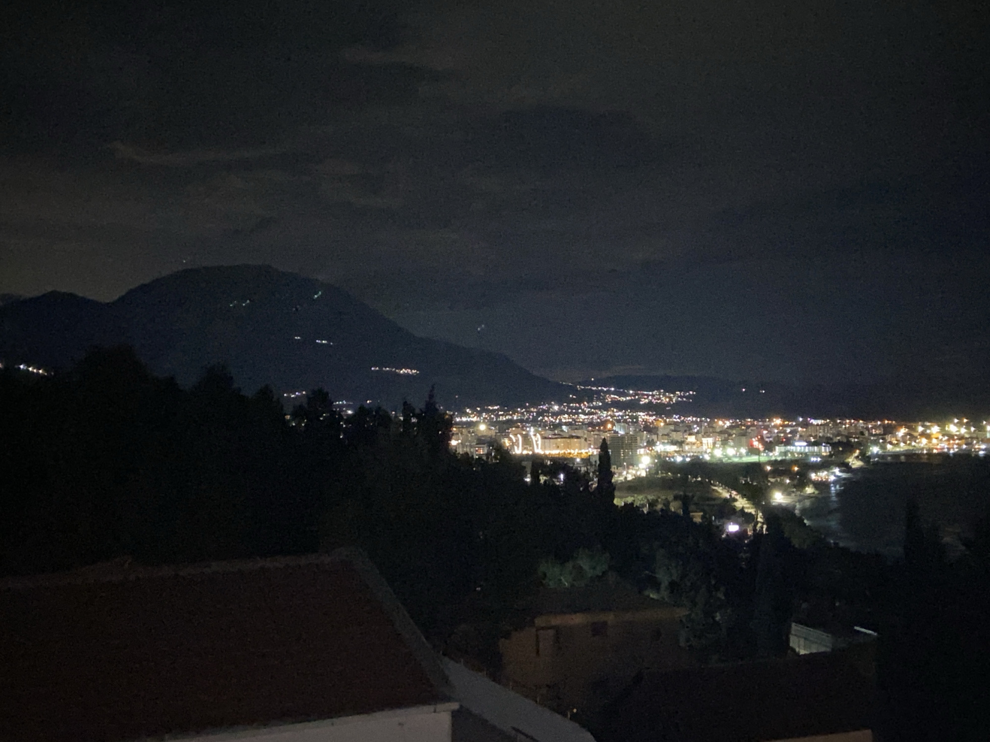 A nighttime shot shows the city lights of Šušanj, Montenegro, from an elevated perspective. The foreground features a rooftop and dark trees, with the city lights extending into the middle ground. A mountain range is visible in the background under a dark, cloudy sky. The city lights indicate a coastal area, likely reflecting off the sea. No people are visible in the scene.