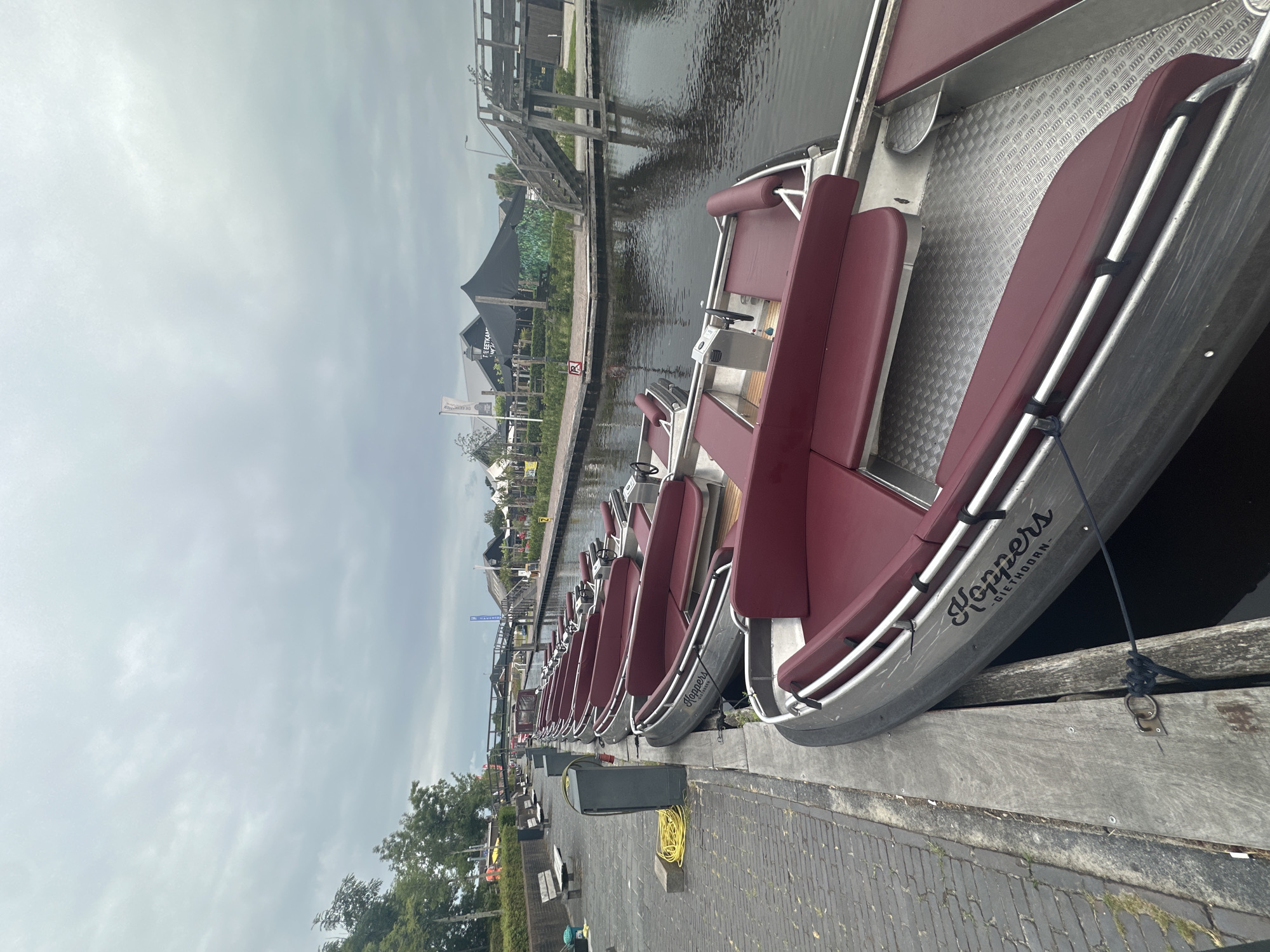 The image depicts a canal in Giethoorn, Netherlands, lined with multiple small, silver motorboats with maroon upholstery. The boats are docked side-by-side along a paved walkway constructed of interlocking bricks and wooden planks. The walkway features benches, a grey waste receptacle, and a coiled yellow electrical cord. In the background, several buildings with dark roofing and awnings are visible, along with a wooden pedestrian bridge spanning the canal. The sky is overcast with grey clouds. The boat in the foreground has the word "Hoppers" and "-GIETHOORN-" written in black script on its side. There are no people visible in the image.