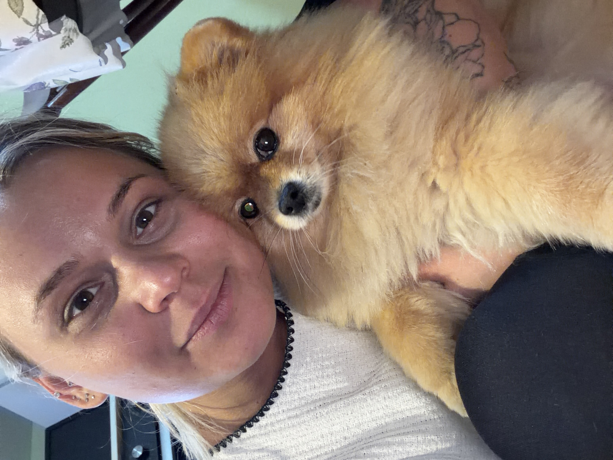 A woman with blonde hair is holding a tan-colored Pomeranian dog. The woman is wearing a white shirt with black trim. The dog has dark eyes and a black nose. The background is a light green wall. The location is not identifiable as Sarapul, Russia, based solely on the image.