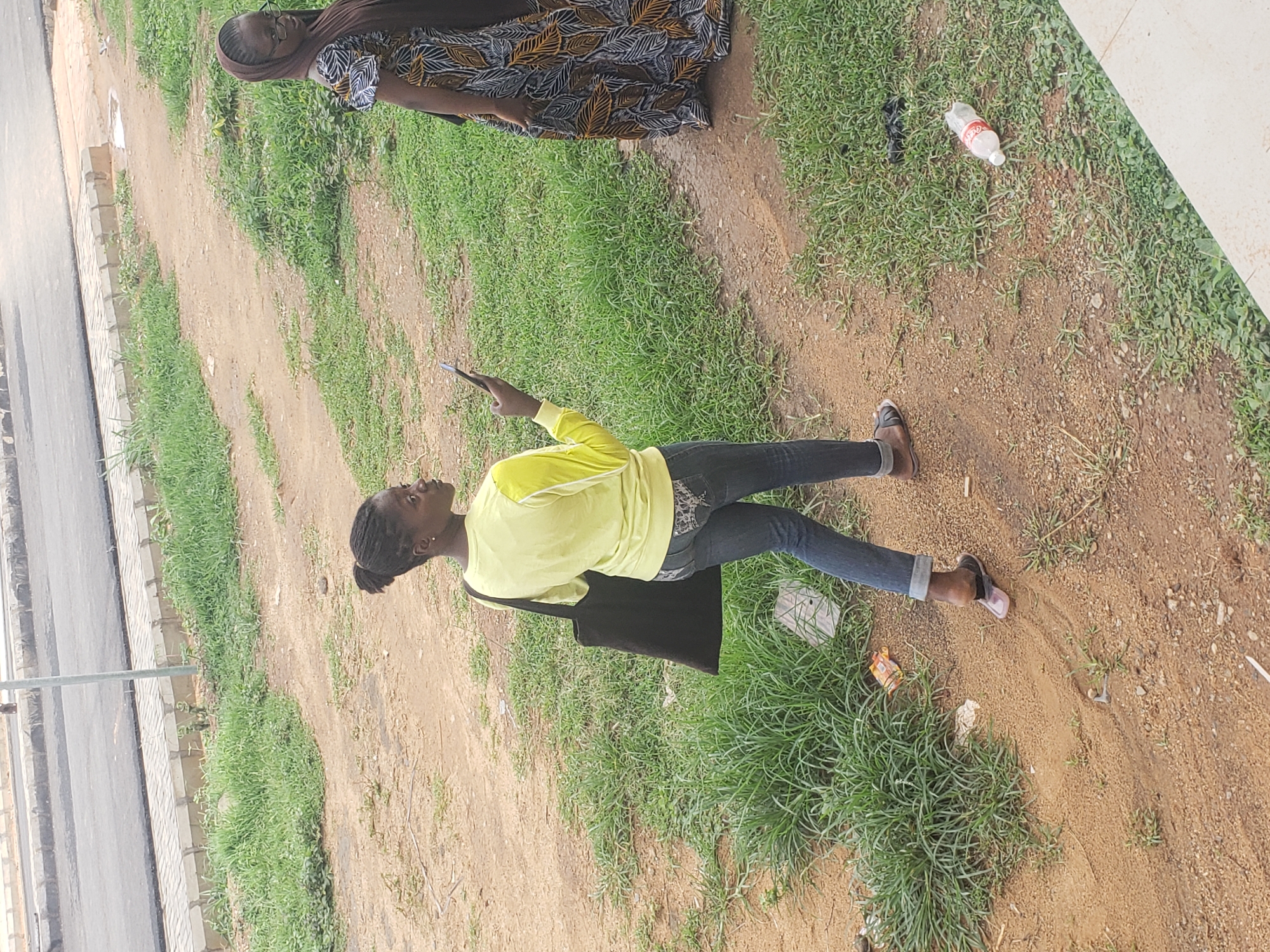 In Gumel, Nigeria, a young Black woman with her hair pulled back in a bun, wearing a bright yellow long-sleeved shirt and dark blue jeans, is walking away from the camera on a dirt path. She is carrying a large black tote bag over her left shoulder and holding a black smartphone in her right hand, looking at it. To her right and further down the path, another Black woman is standing, wearing a brown hijab and a patterned dress with black, white, and yellow designs. The environment is characterized by a dirt path cutting through an area of sparse grass and vegetation, with a paved road and sidewalk visible in the background. There is some litter on the ground, including a discarded plastic bottle and other debris.